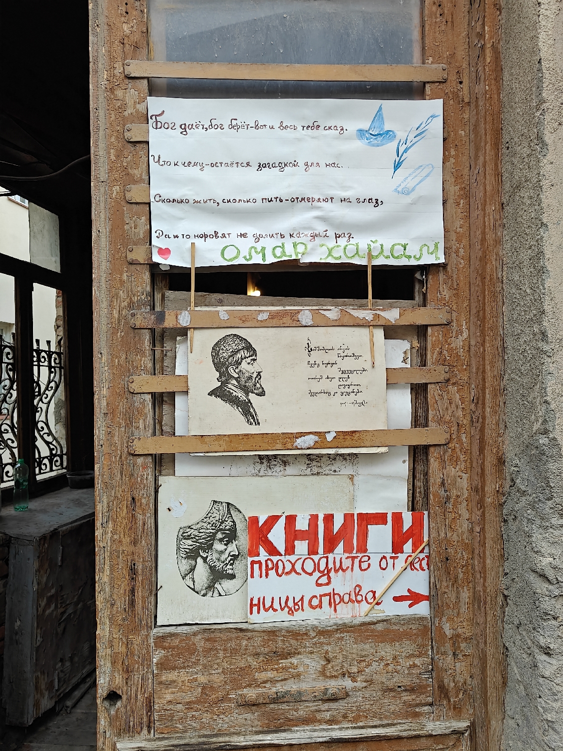 The image captures a weathered, peeling wooden door or window frame, likely part of an old building, adorned with several handwritten and hand-drawn signs. The scene is illuminated by daylight. Through an opening to the left, intricate metalwork and a distant green bottle can be seen, hinting at an alleyway or courtyard in Tbilisi, Georgia.
Three signs are prominently displayed on the wooden frame. The topmost sign is a white sheet of paper featuring a quote in Russian attributed to "ОМАР ХАЙЯМ" (Omar Khayyam). The text translates to: "God gives, God takes - that's all there is to say. What for what - remains a mystery to us. How much to live, how much to drink - they measure by eye, and they try not to pour enough each time." Alongside the text are small illustrations of a blue pointy hat, a feather, and a rolled scroll.
Below this, a second, aged paper sign is visible. On its left, there's a hand-drawn black-and-white portrait of a bearded man wearing a distinctive cap, likely Omar Khayyam himself. The right side of this sign contains text in Georgian, which includes phrases like "No one will challenge/compete with us. Even the dead will laugh."
The lowest sign is another white paper, with a third hand-drawn portrait on the left, similar in style to the one above, depicting another historical figure. The right side features large, bold red letters spelling "КНИГИ" (BOOKS), followed by smaller red text directing: "проходите от лестницы справа" (go from the staircase on the right), accompanied by a red arrow pointing right, indicating direction to a bookstore or a book-related area.
The overall impression is one of a charming, possibly bohemian, establishment with a philosophical and literary bent, inviting passersby with wisdom and clear directions.