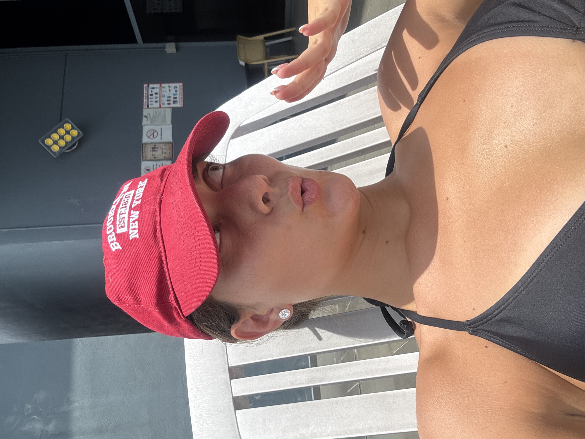 A young woman is depicted reclining outdoors on a white slatted lounge chair under bright daylight, likely in Kuala Lumpur, Malaysia. She is wearing a black bikini top and a red baseball cap, with her gaze directed upwards and her mouth slightly open, suggesting relaxation or an expression of warmth. Her left ear is visible, adorned with a small, circular stud earring. The cap prominently displays white text that reads "BROOKLYN EST.1837 NEW YORK".
Part of another person's hand, with neatly manicured fingernails, is visible reaching down towards her face from the top right. The harsh sunlight casts strong shadows, indicating a sunny day, likely midday or early afternoon.
In the background, a dark gray wall features a black light fixture with nine prominent yellow circular lights. Below this, several framed signs or notices are mounted on the wall, though their specific content is not clearly discernible. The overall scene suggests a leisure activity in an urban outdoor setting, such as a rooftop deck or poolside area.
