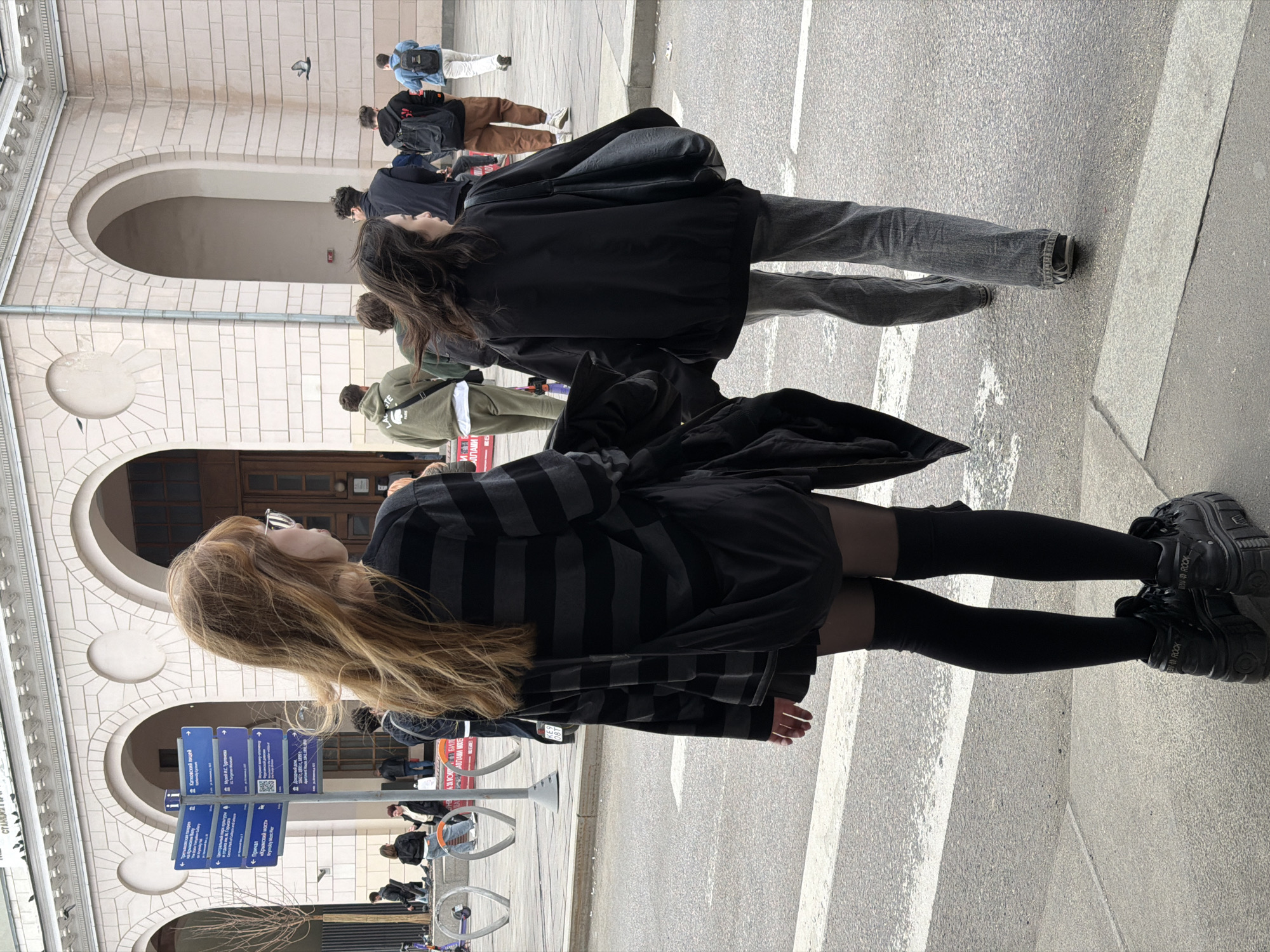 The image depicts a group of people on a pedestrian crosswalk in Moscow, Russia. The backdrop features an ornate, light-colored building with arched windows and decorative circular elements. Street signs with Cyrillic text are visible on a post near the crosswalk.
In the foreground, a person with long, light-colored hair is facing left, wearing a striped dark shirt, black tights, and platform black shoes. Behind her, another person with dark hair and a black jacket is walking. The background includes other pedestrians and elements of the urban landscape. The focus is on the street and its immediate surroundings.