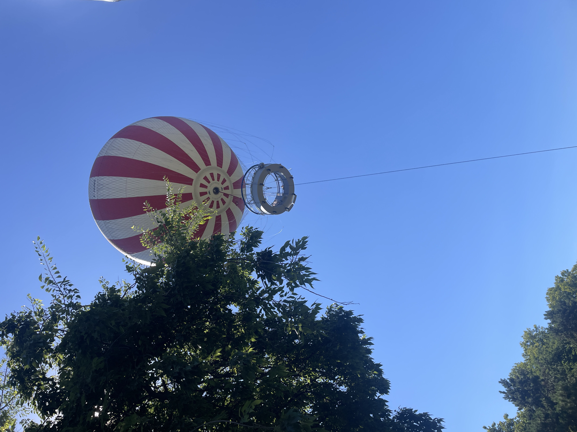The image shows a tethered hot air balloon against a clear blue sky. The balloon is red and white striped. A circular gondola is suspended below the balloon. The lower portion of the image features the tops of trees. The hot air balloon is located at the Budapest Eye observation wheel attraction in Budapest, Hungary.