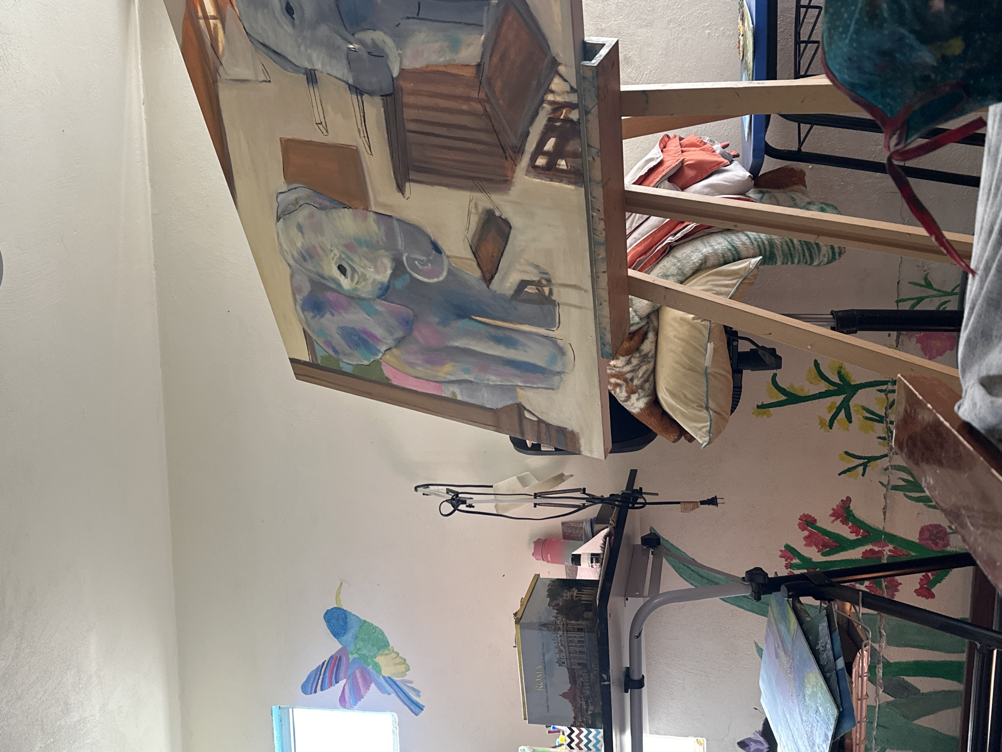 The image depicts an indoor space with an unfinished painting of two figures, possibly elephants, on an easel. The painting shows the subjects in a simplified, abstracted style with shades of gray, blue, and pink. The setting appears to be an artist's studio or workspace, with various art supplies, cushions, and other items scattered around. The walls feature hand-painted floral designs and a whimsical bird near a window. The city and country location are Puebla, Mexico.