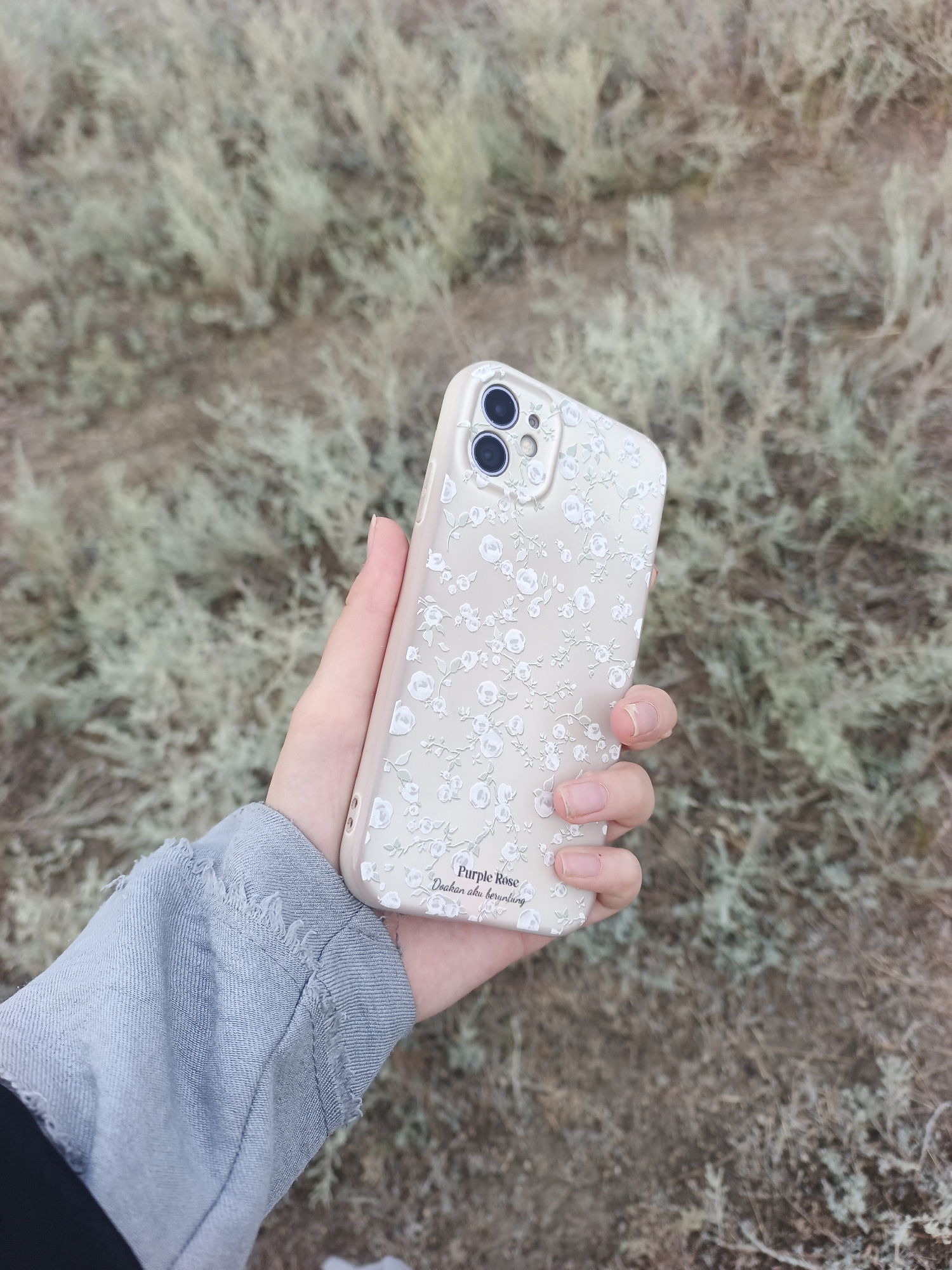 A person's hand, wearing a light blue denim jacket with frayed cuffs, holds a light beige phone case with a delicate white floral pattern. The phone case has two prominent black camera lenses on the top left. At the bottom of the case, there is text that reads "Purple Rose" and "Doakan aku beruntung". The background is a blurred landscape of dry, light-colored grass and earth. The scene appears to be outdoors in Varenikovskoe, Russia, though the exact location within the city is not specified.