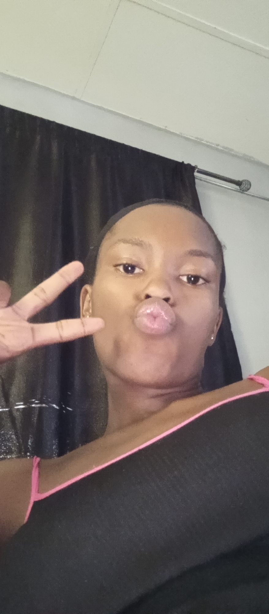 A young woman is taking a selfie indoors, with a close-up view focusing on her face and upper body. She is making a kissing face and holding up a peace sign with her left hand. She is wearing a black top with pink straps. In the background, there is a dark curtain and a section of a light-colored wall with a metal bar. The location is Unknown, Unknown.