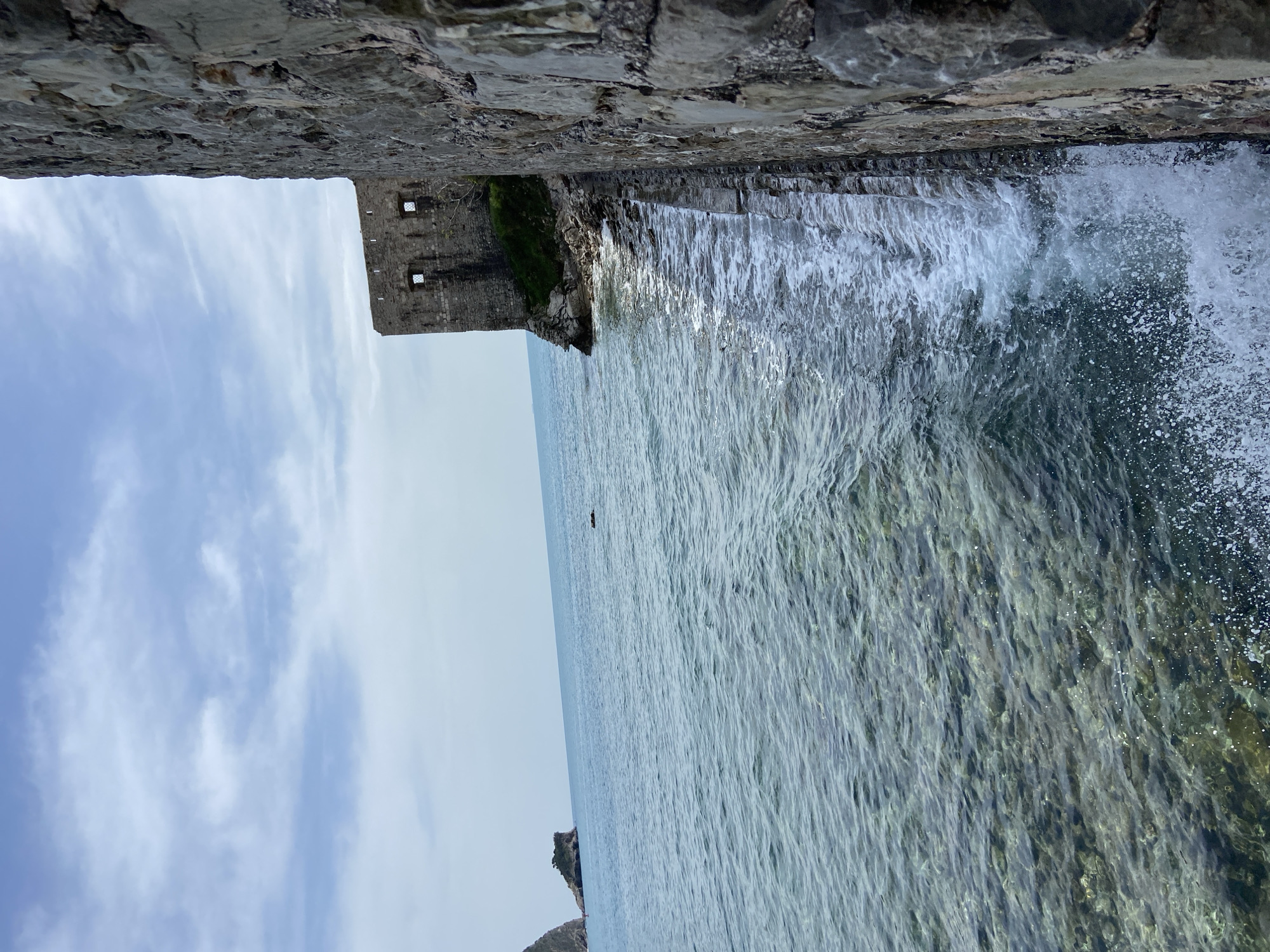 The image presents a scenic coastal view, likely from atop or beside an ancient stone structure. In the immediate foreground, a rugged, textured stone wall occupies the top third of the frame, hinting that the viewer is looking out from a fortification or high vantage point.
Below the wall, the sea dominates the scene. On the right side, foamy white waves crash against the shore or rocks, creating splashes and showing turbulent, clear water revealing submerged rocks. Further out, the water transitions to a calmer, serene expanse of blue-green, displaying subtle ripples under a bright sky. A small, dark, elongated object is visible floating in the distance on the calm water, its exact nature unclear but possibly a distant boat or person.
In the mid-ground, to the left, a historic stone building or ruin stands prominently on a rocky headland, featuring several small, rectangular openings that resemble windows or battlements. The structure appears weathered, with patches of green vegetation clinging to the rocks around its base. Another smaller, dark rocky outcrop is visible further left and closer to the water.
The sky, filling the left portion of the background, is light blue with scattered, wispy white clouds, indicating a bright daytime setting. There is no discernible text in the image. The scene conveys a sense of historical presence intertwined with natural beauty, characteristic of coastal regions like Budva, Montenegro.