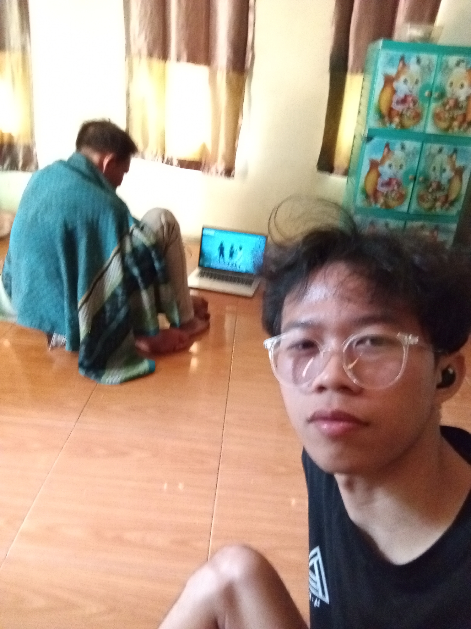 The image depicts an indoor scene with two individuals and a laptop. The person in the foreground is facing the camera, wearing glasses and a black t-shirt, with earbuds in. Behind them, another person is seated on the floor, covered by a teal blanket, facing away from the camera and looking at a laptop which displays figures. The room has light-colored tiled flooring and curtains in the background. To the right, a blue cabinet with cartoon fox designs is visible. The overall environment suggests a domestic setting. This scene could be located within a residential area in Subang, Indonesia, as the cultural context of the cabinet and typical home furnishings can be associated with Indonesian households.