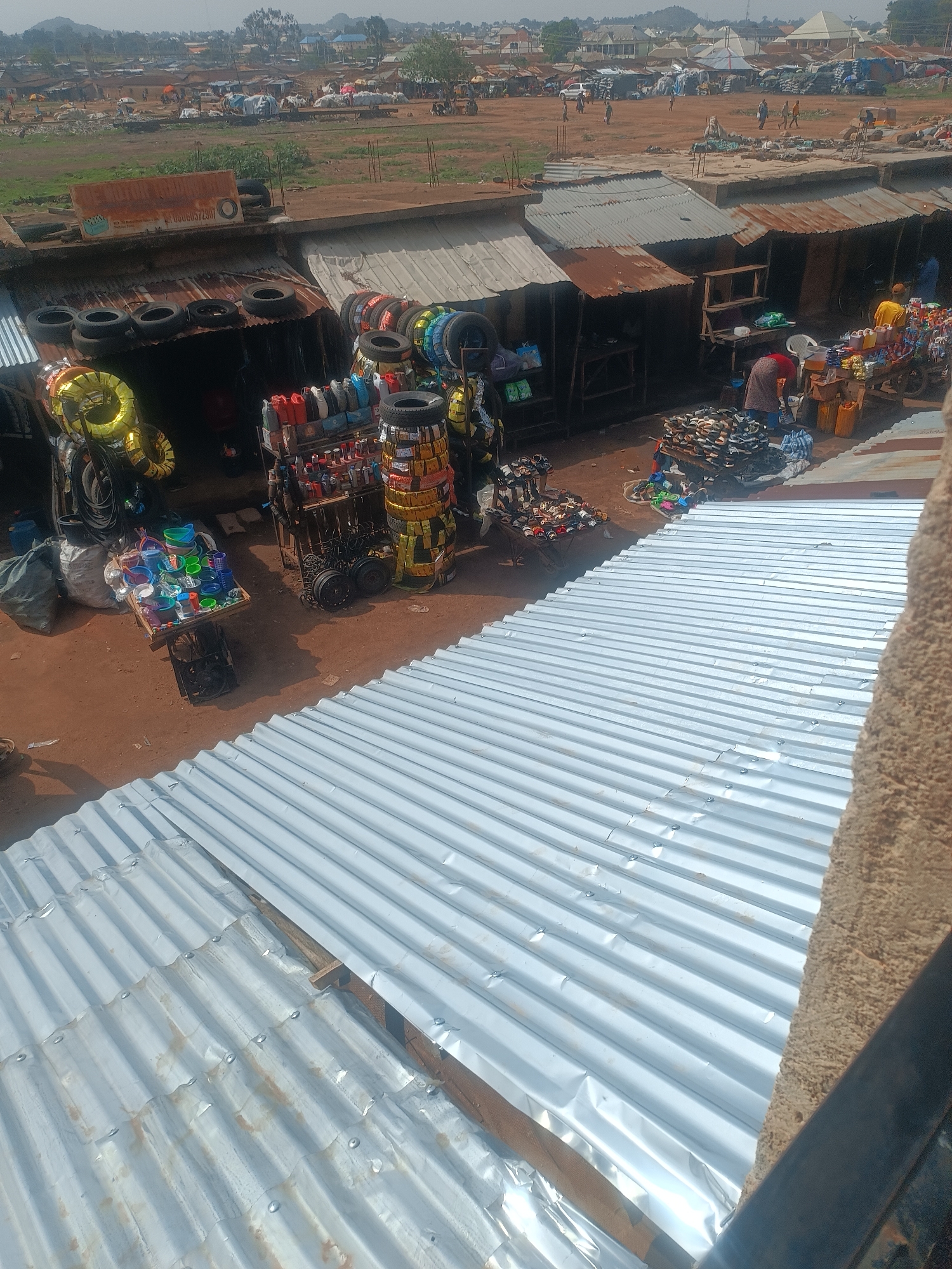 The image depicts an outdoor marketplace scene in Bukuru, Nigeria, viewed from an elevated perspective, likely from a building. The foreground is dominated by corrugated metal roofs. Beyond the roofs are a series of market stalls selling various goods, including tires, motor oil, sandals, and plastic containers. People are seen browsing and interacting with the vendors at the stalls. In the background is an open, dirt-covered field where other people are walking around. Buildings and trees are also seen in the distance. The overall environment suggests a bustling, active marketplace.