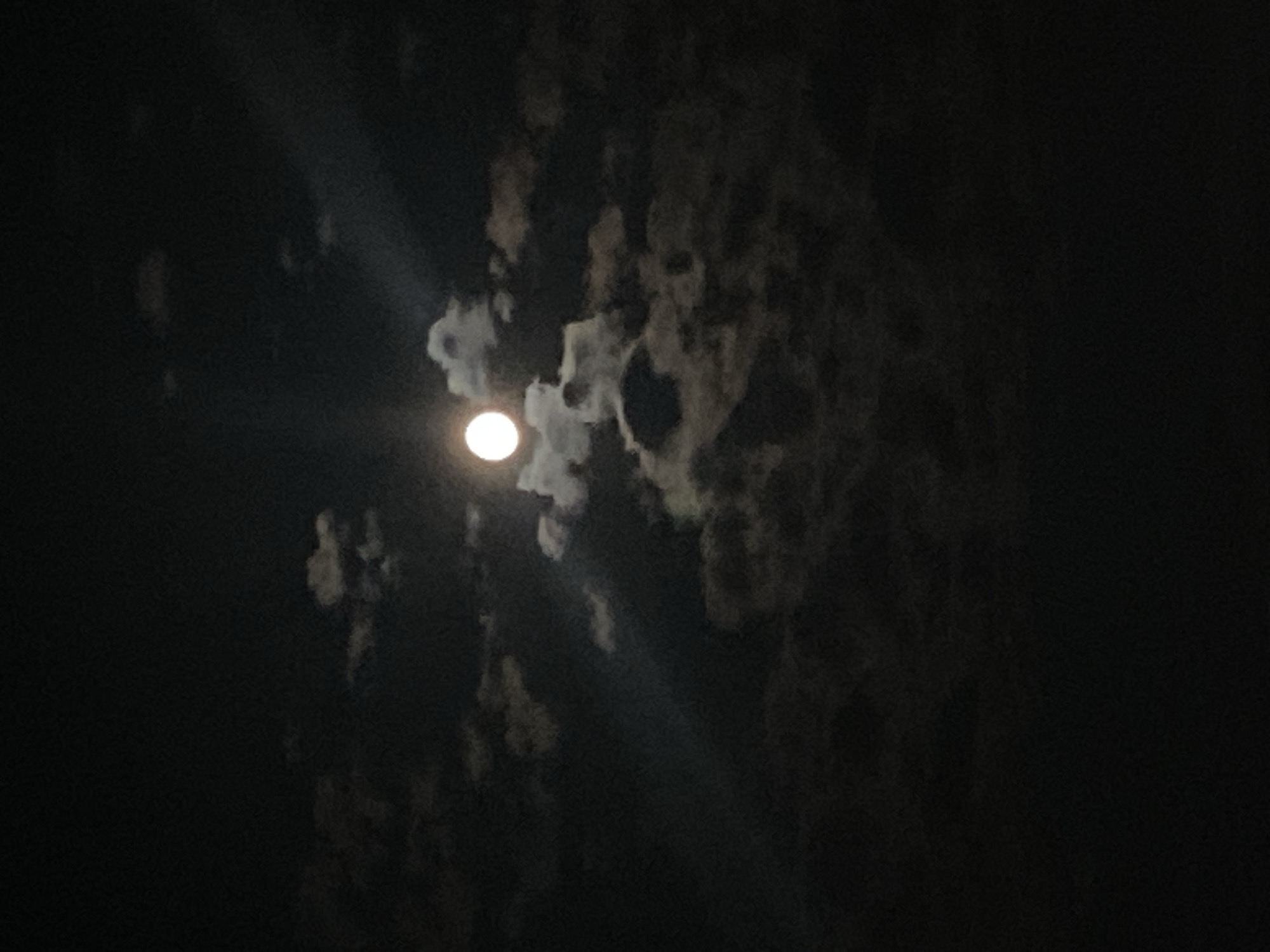 A full moon is visible in a dark, cloudy night sky. The moon is a bright, white orb surrounded by wispy clouds. Beams of light emanate from the moon, piercing through the clouds. The overall scene is dark and atmospheric. The image does not contain any identifiable landmarks or features that would allow for specific location identification within the city of Iași, Romania. There are no people or discernible objects in the scene other than the moon and clouds.