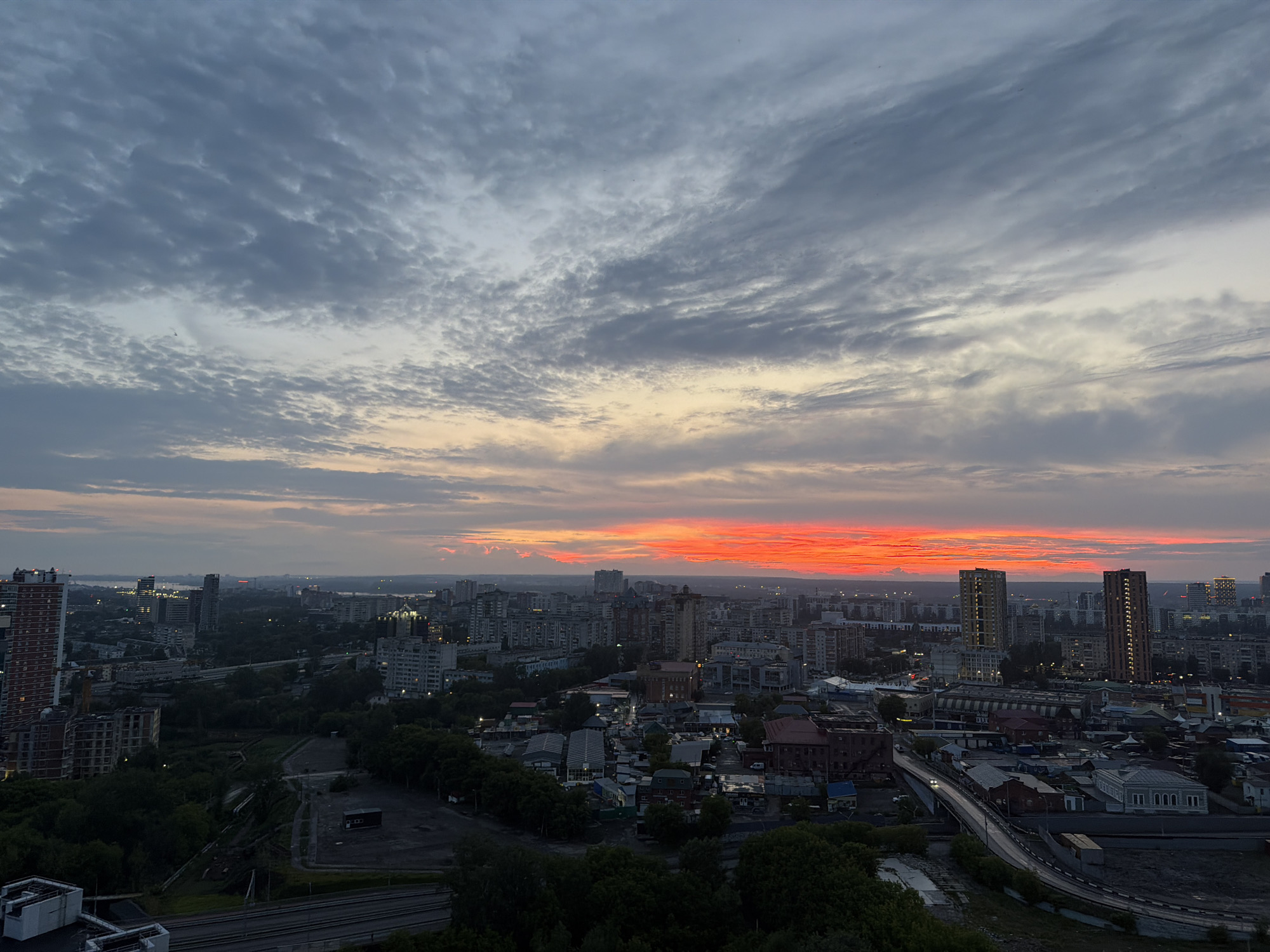 This is a high-angle, wide-shot image of a cityscape at dusk, likely in the city of Perm, Russia, given the context of similar images. The sky is filled with dramatic clouds, with a vibrant band of orange and red light near the horizon, suggesting a sunset or sunrise. Below the sky, a dense urban environment stretches out, characterized by a mix of residential and commercial buildings of varying heights. Several tall, modern buildings stand out amongst older, lower-rise structures. Greenery, in the form of trees, is visible in patches throughout the city, particularly in the foreground and mid-ground, suggesting parks or tree-lined streets. The lighting is dim, with some artificial lights starting to appear in the buildings and along roads, indicating the transition from daylight to night. The overall scene depicts a city at the cusp of evening, bathed in the warm glow of the setting or rising sun, with a sense of urban scale and activity. No people are distinctly visible in the image.