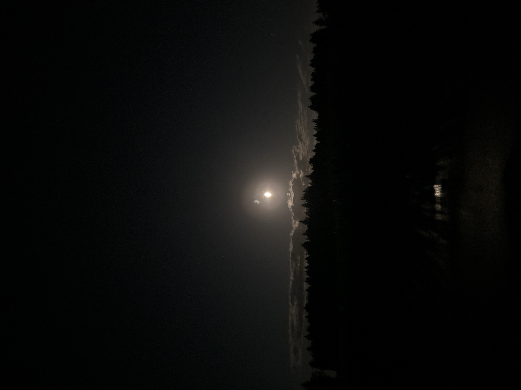 A full moon illuminates a dark sky over a silhouetted forest line. The moon is partially obscured by thin, wispy clouds, creating a soft glow. Below the tree line, reflections of the moonlight are visible on what appears to be a body of water or a wet surface, casting elongated shadows. The foreground is entirely dark, obscuring any details of the ground or what lies beyond the reflections. The scene is devoid of human presence or activity. The image was captured in the vicinity of Krautscheid, Germany.