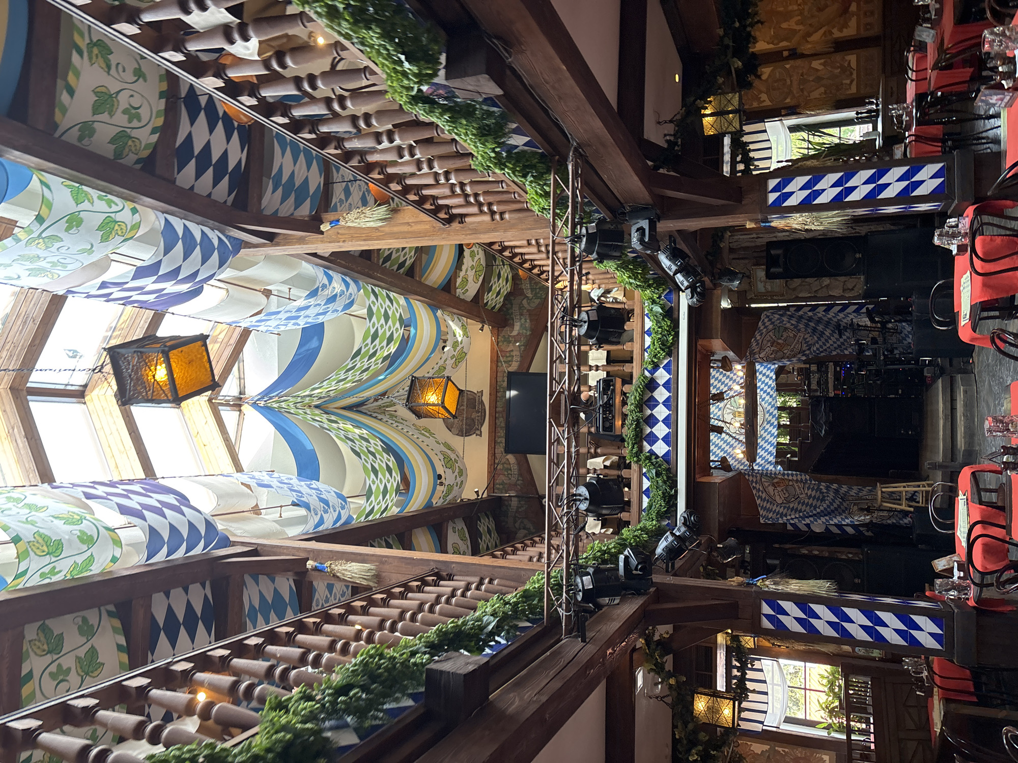 This image captures the festive and elaborately decorated interior of a Bavarian-themed restaurant or hall, located in Saint Petersburg, Russia. The view is from an elevated position, looking down into the main space.
The scene is dominated by robust wooden beams and railings, which are extensively adorned with lush green garlands, adding a natural touch to the rustic architecture. The ceiling is a prominent feature, draped with numerous fabric panels displaying a variety of patterns: classic blue and white diamonds, evocative of the Bavarian flag, interspersed with sections featuring green and white stylized hops or vine motifs. Two decorative, dark metal lanterns with amber-colored glass panels hang from the ceiling, casting a warm, inviting light.
Towards the right side of the image, a dining area is visible with tables uniformly set with red tablecloths and chairs, some featuring matching red chair covers. Glasses are arranged on the tables. In the background and center, a designated performance area or stage is set up, complete with large black speakers, an array of spotlights, and other technical equipment, suggesting live entertainment. This area also incorporates blue and white diamond-patterned tiles on its supporting structures and a banner with a blue and white checkered pattern, displaying a crest or logo with text that is not discernible. Small bundles of dried wheat or straw, tied with blue ribbons, serve as charming decorative accents on the railings. A small television screen is mounted on one of the wooden beams.
The space is illuminated by a combination of natural light filtering through what appear to be skylights and a window on the far right, along with artificial light from the lanterns and spotlights. No people are present, indicating a moment before or after an event, or during an off-peak hour. The overall atmosphere is prepared and inviting, ready for guests and activities.
