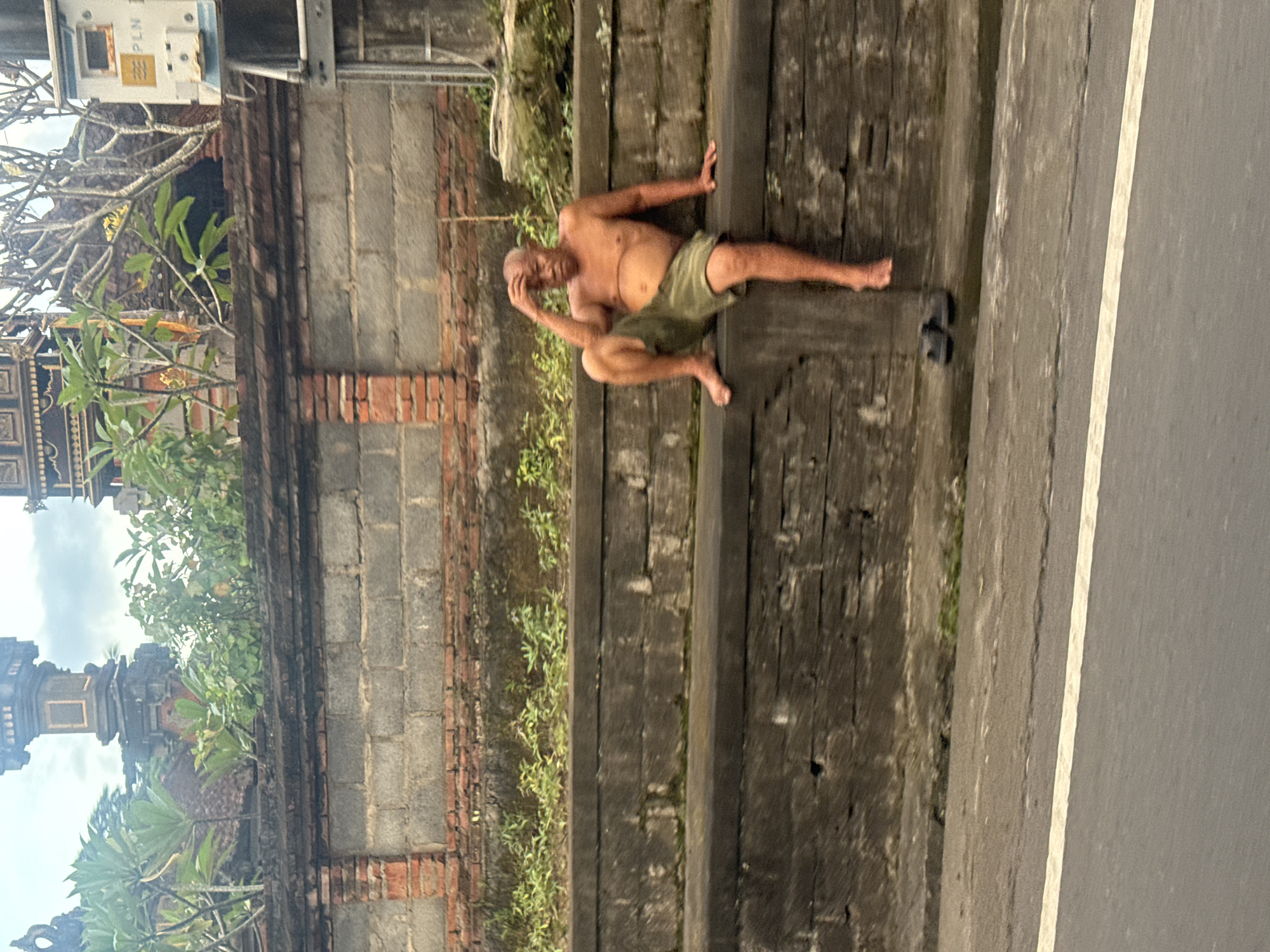 The image shows an older man in shorts sitting on a stone wall next to a road in Sukawati, Indonesia. He is shirtless, balding, and appears to be resting his head in his hand. A pair of sandals sit on the wall near his feet. The wall itself is made of dark stone. A brick wall with vegetation growing on it runs parallel to the stone wall. A building with traditional Balinese architecture is visible in the background.
