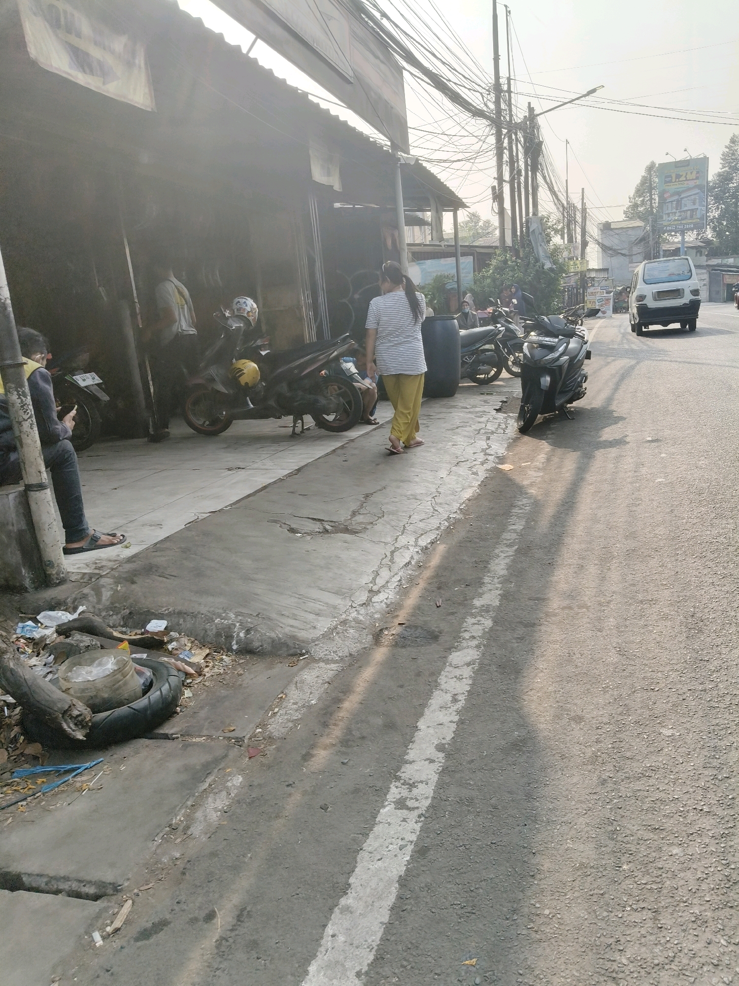 The image depicts a street scene in South Tangerang, Indonesia, on a sunny day. On the left, a row of buildings with corrugated metal roofs and awnings line the street. In front of these buildings, several motorcycles are parked. A person is seated on a low wall, looking at their phone, while another person walks away from the camera. A large black plastic barrel is also visible. The sidewalk area is paved and appears somewhat worn, with debris and a discarded tire scattered near the edge. The street itself is asphalt, with a white lane marker visible. Power lines crisscross overhead, supported by utility poles. In the background, a white van is driving on the road, and a billboard is visible further down the street. The overall impression is of a busy, urban environment.