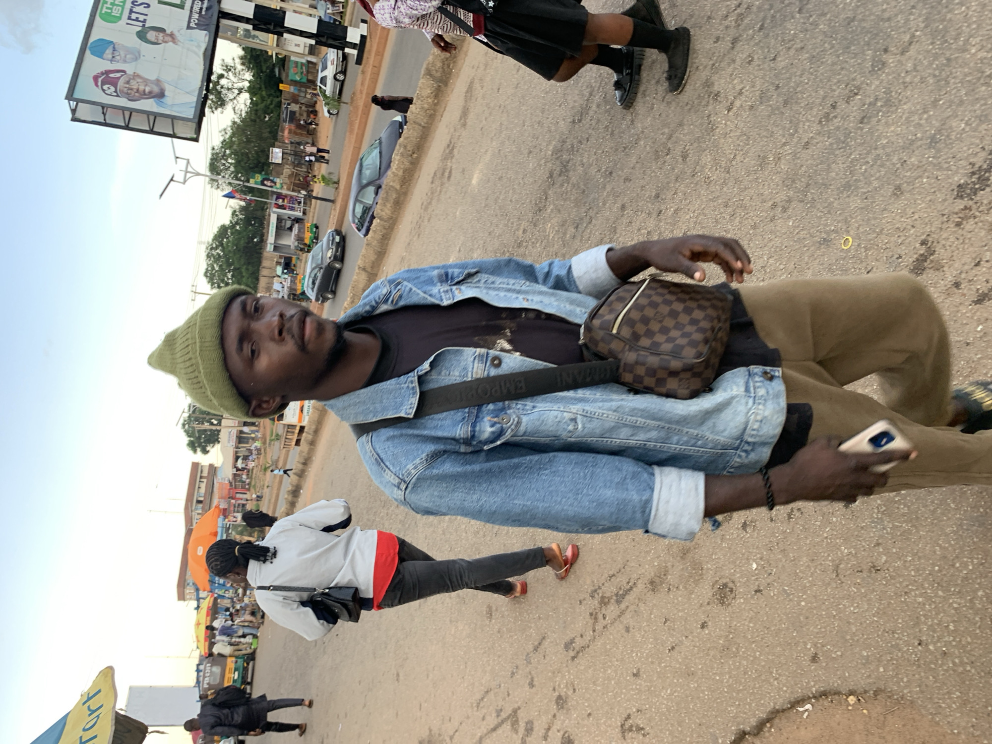 The image captures a street scene in Jos, Nigeria, at dusk. In the foreground, a young man walks towards the camera, wearing a green beanie, a denim jacket, and tan trousers. He carries a brown and checkered crossbody bag and holds a mobile phone in his right hand. Behind him, another person, likely a woman, walks away from the camera, dressed in dark trousers, a red top, and a white jacket, with a black handbag slung over her shoulder. Further in the background, several other individuals are visible, along with parked vehicles, market stalls, and a large billboard displaying political figures. The environment appears to be an urban setting with a paved road and sidewalk, suggesting a bustling commercial or residential area. The overall lighting indicates the soft glow of late afternoon or early evening.