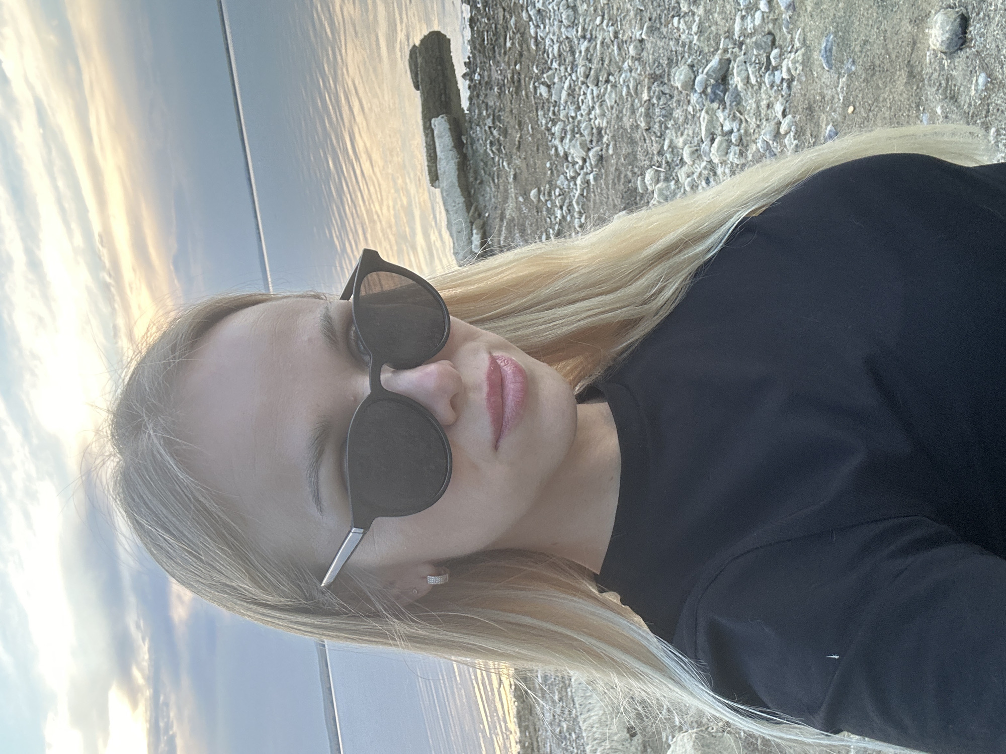 A young woman with blonde hair and sunglasses takes a selfie on a rocky shore. The woman is wearing a black t-shirt. The background features a body of water and a cloudy sky. The location is the shore of Lake Gidroelektrostantsii in the city of Energetik, Russia.