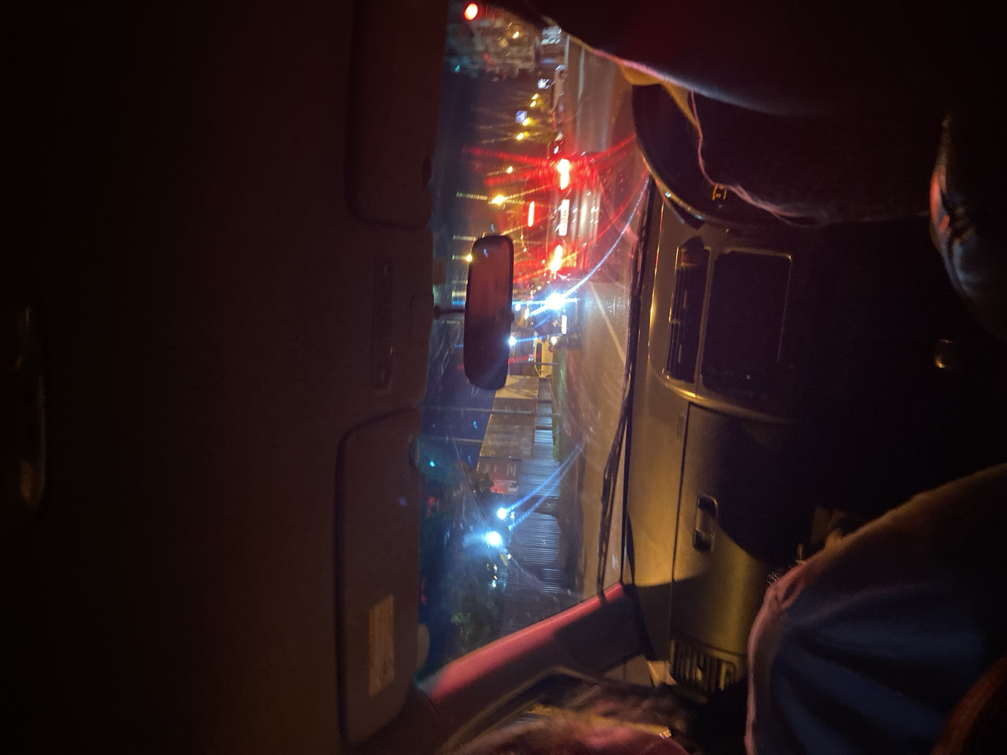 The image captures the interior of a car at night, looking out through the windshield. The scene depicts a street with moderate traffic. Several cars are visible, with their headlights and taillights illuminated, creating streaks of light due to the long exposure or wet conditions on the road. The environment appears to be urban, with buildings and streetlights in the background, although specific details are obscured by darkness and light reflections. Two individuals are partially visible inside the car, one in the passenger seat on the left and another in the driver's seat on the right, their faces and bodies mostly in shadow. The dashboard of the car is dimly lit, with some reflections from the external lights. The overall impression is a nighttime journey through a city street, possibly in rainy or wet conditions, given the light reflections and streaks.