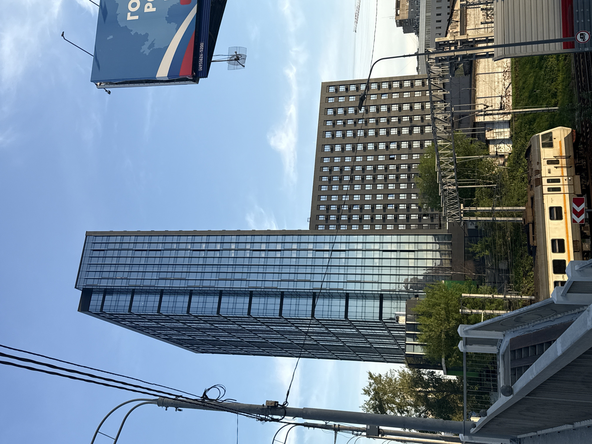 The image presents an upward view of the urban landscape in Moscow, Russia, under a clear, blue sky with scattered clouds. Two buildings dominate the scene: a tall, modern skyscraper with reflective glass panels and a taller, older, rectangular building with regularly spaced windows. In the foreground, utility poles and power lines intersect the view, adding to the complexity of the urban environment. A portion of a billboard with the Russian flag is visible at the top left corner. Additionally, a train and railway structure can be seen in the mid-ground, adding another layer to the urban scene.