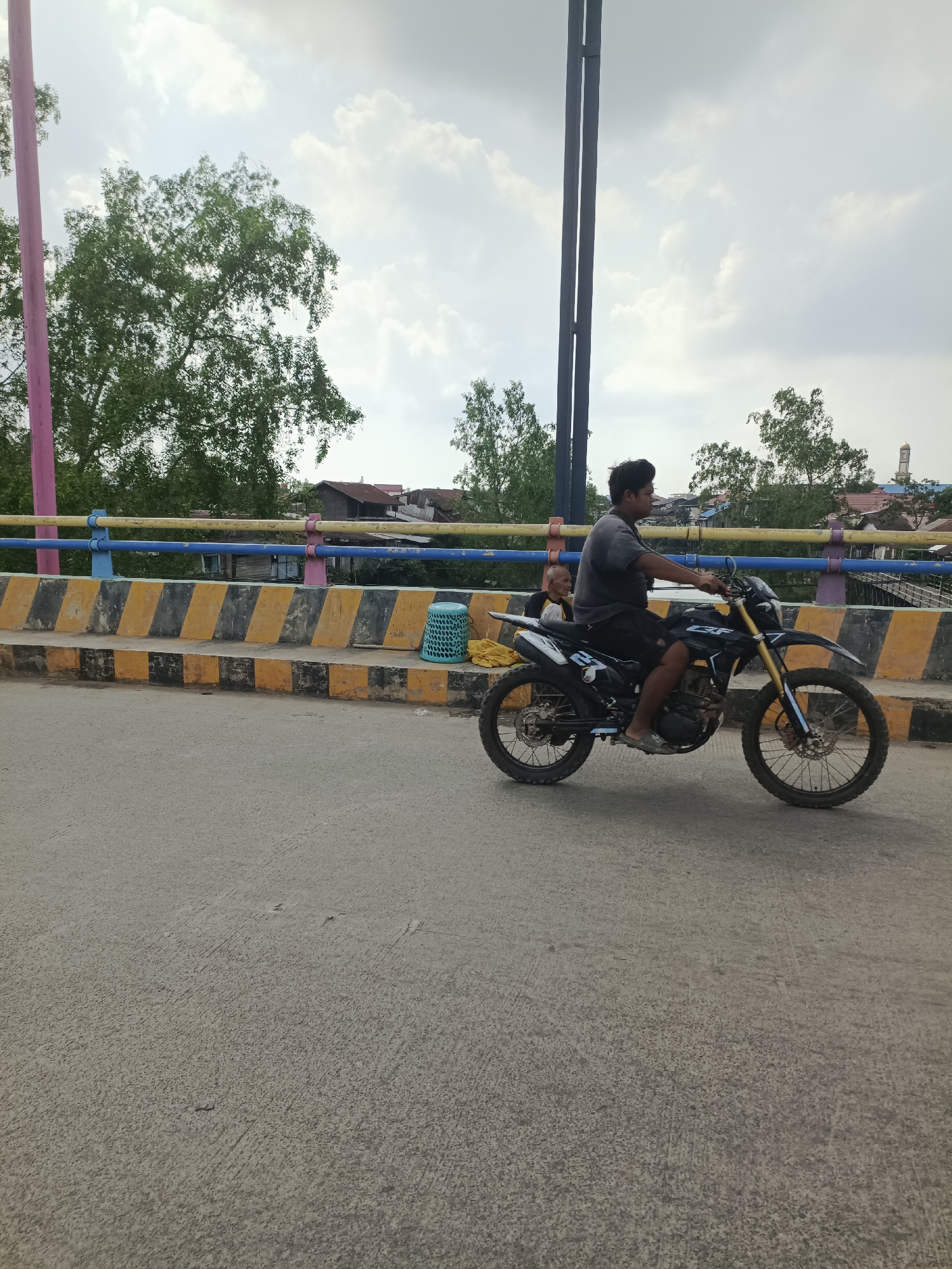 A medium shot shows a man riding a black dirt bike on a paved road with a bridge in the background. The man is wearing a dark t-shirt, dark shorts, and flip-flops. He is looking to his left. Behind him, on the bridge, a child is sitting next to a blue plastic basket. The bridge has yellow and black striped barriers, and a blue and yellow railing. Trees and buildings are visible on the other side of the bridge. The sky is cloudy. This scene is located in Sangatta, Indonesia.