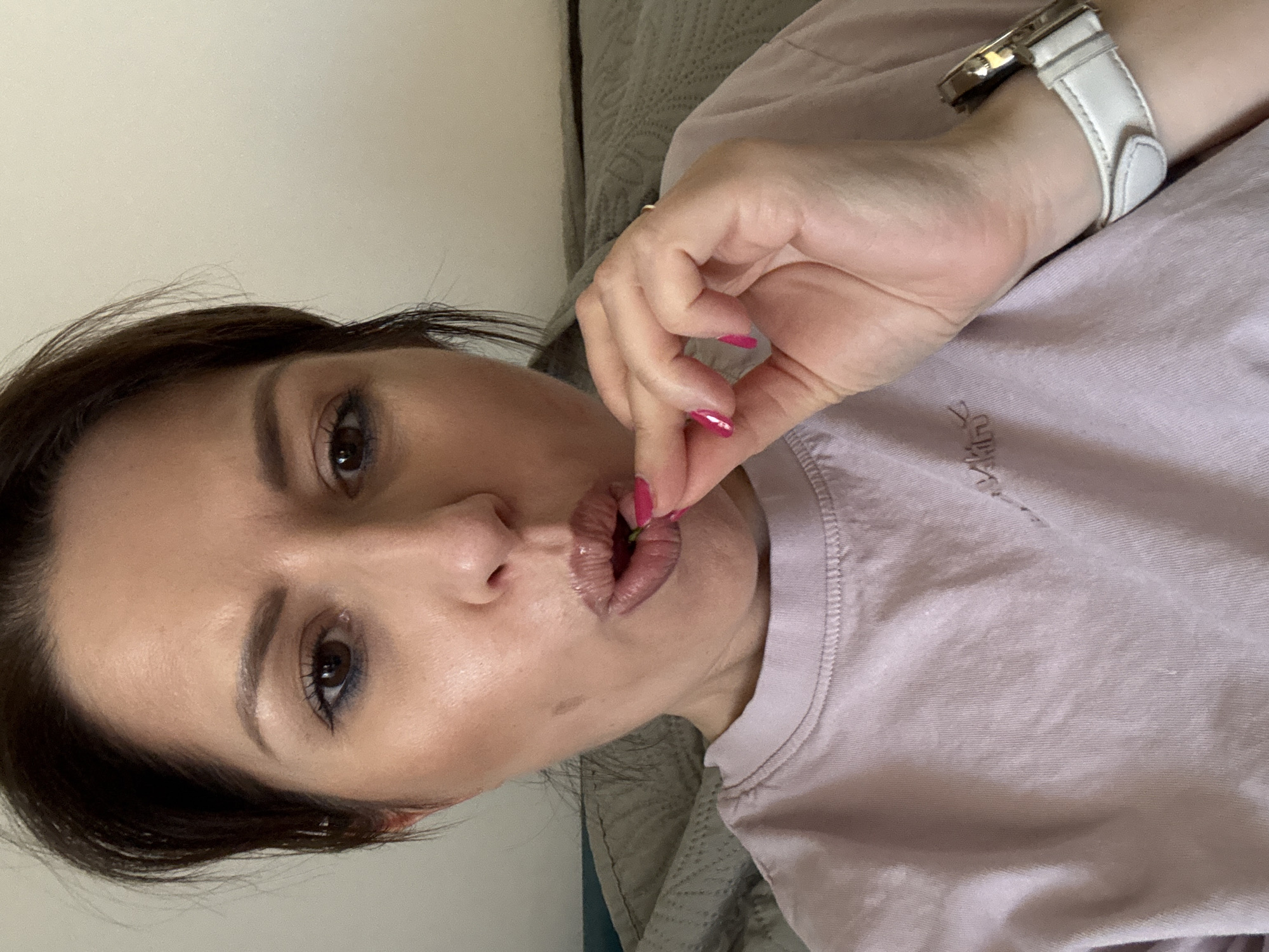 A woman with dark hair and light skin is holding a small, red object to her lips. She is wearing a light pink t-shirt and a white watch. The background is blurred, but appears to be a bed with gray bedding. The photo does not provide enough information to determine the location in Sofia, Bulgaria.