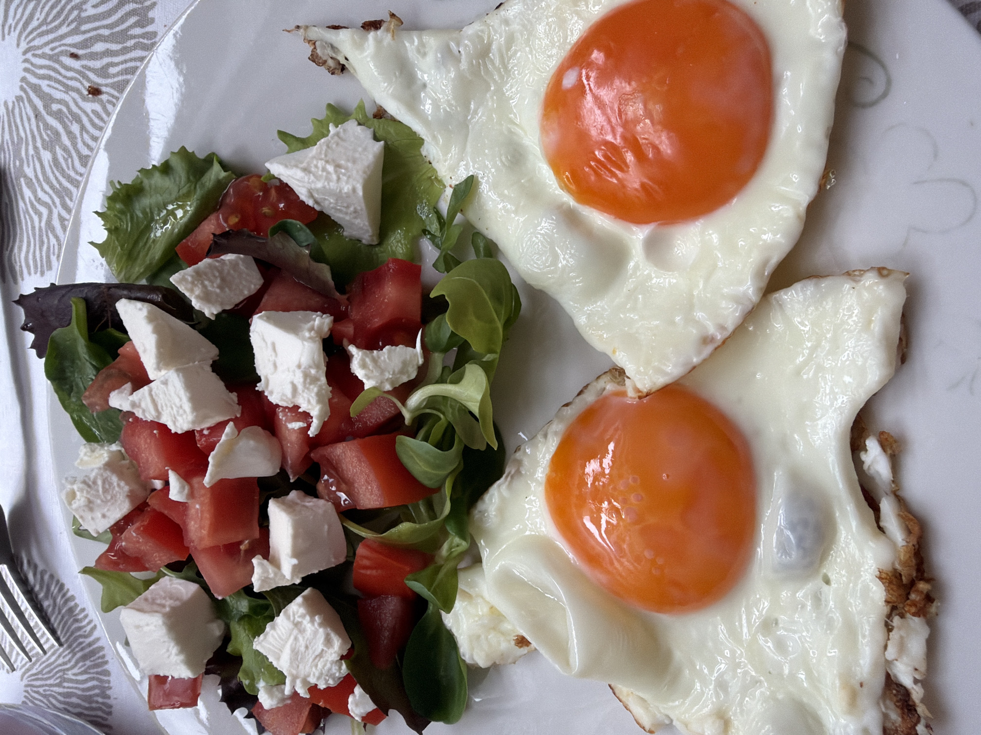 A top-down view of a white plate displaying a meal. The meal consists of two fried eggs with bright orange yolks and slightly browned edges. Adjacent to the eggs is a salad of mixed greens, diced tomatoes, and cubes of white cheese. A fork is visible on the left side of the plate. The plate rests on a surface with a white and gray patterned tablecloth. The location is not discernible from the image alone, and information about Budva, Montenegro is not present in the photograph.