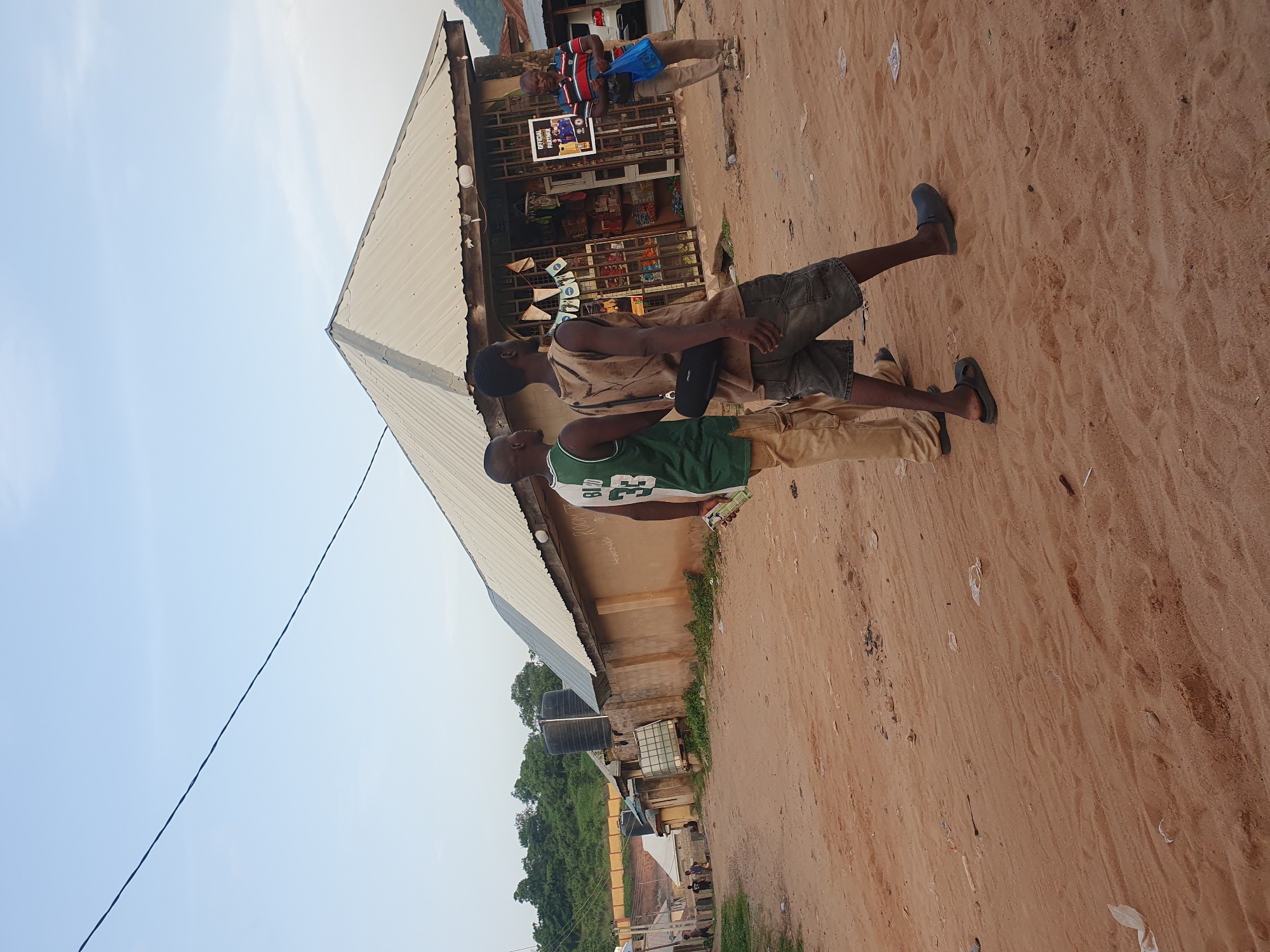 This is a medium shot, taken at eye-level, of a street scene in Lokoja, Nigeria. The ground is sandy and uneven, with scattered debris such as plastic bags and paper. On the right side of the frame, a small, open-fronted shop with a corrugated metal roof is visible. The shop is stocked with various items on shelves. In front of the shop, a man wearing a striped t-shirt and holding a blue plastic bag stands facing left.
In the mid-ground, two young men are walking away from the camera, towards the shop. The man on the left wears a green basketball jersey with the number 33 and the name "BIRD" on it, along with khaki pants and dark sandals. The man on the right wears a tan tank top with a dark bag slung over his shoulder, denim shorts, and dark clogs.
In the background, several low-lying buildings with similar corrugated metal roofs line the street. Water tanks are visible on some of the buildings. Lush green trees and vegetation cover the hillside behind the buildings. The sky is clear and pale blue, with a power line stretching across the upper left quadrant of the image. The lighting suggests it is either early morning or late afternoon.