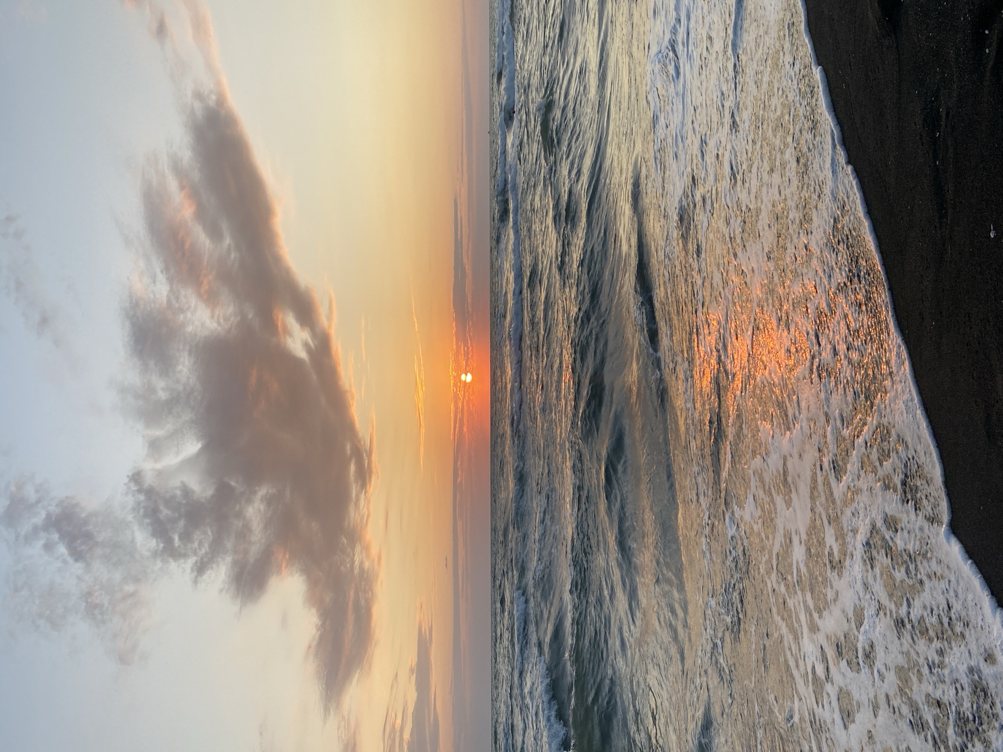 The image depicts a serene sunset over the Black Sea in Ureki, Georgia. The sun, a bright orange orb, is partially obscured by the horizon, casting a warm, golden glow across the water and sky. Wispy clouds, tinged with pink and orange, drift across the pale blue sky. The waves of the sea are a deep teal, with white foam cresting as they approach the shore. The beach in the foreground is composed of dark, coarse sand, with a line of white foam marking the edge of the water. No people or objects are visible in the scene, contributing to the tranquil and natural ambiance.