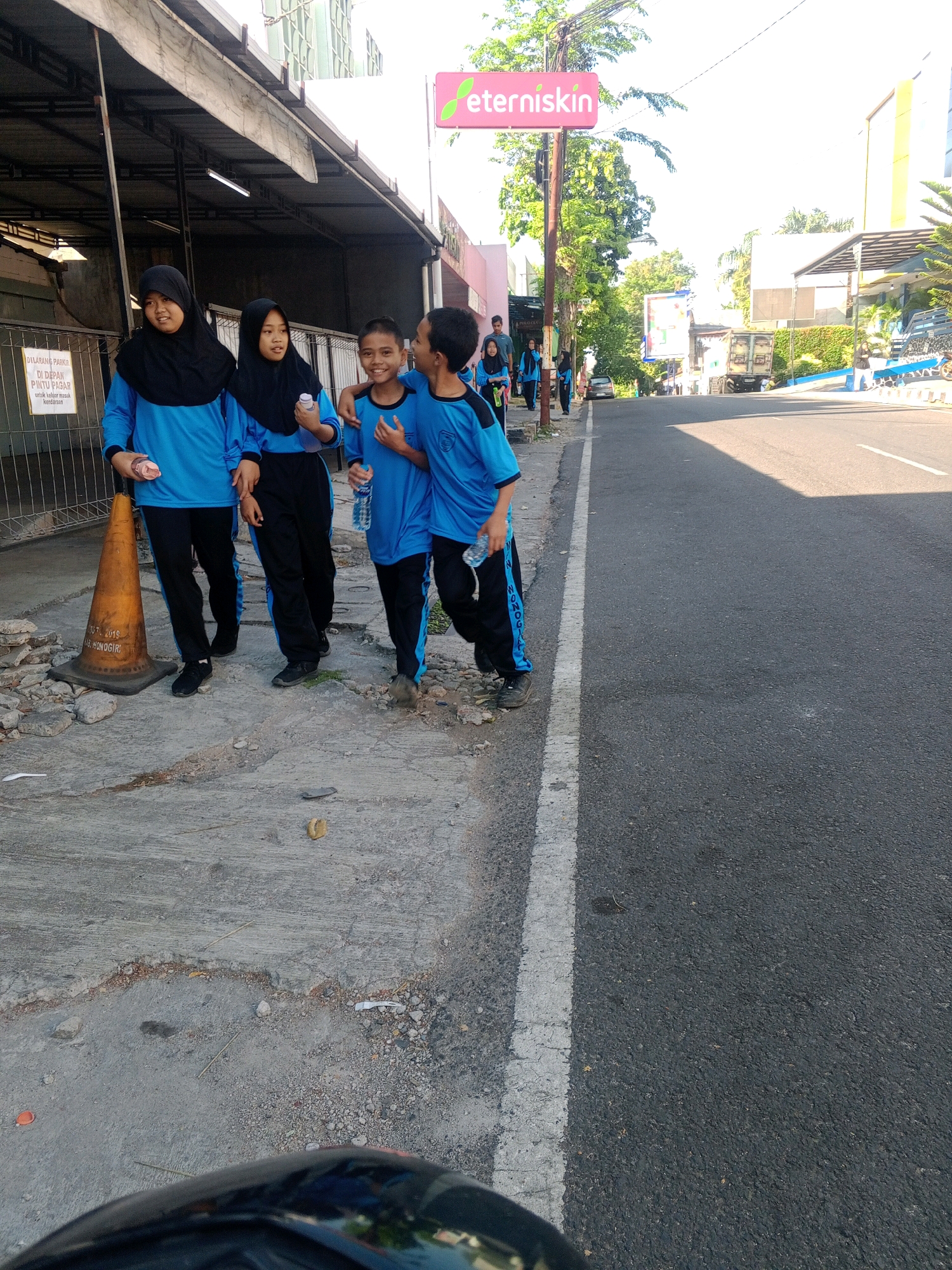 A group of four young people, two females and two males, are walking along a sidewalk adjacent to a street in the city of Wonogiri, Indonesia. The two females on the left are wearing blue shirts, black pants, and headscarves. The two males on the right are wearing matching blue shirts with black accents and black pants. They appear to be in school uniforms. The males have their arms around each other's shoulders. The females are walking slightly ahead of the males.
The sidewalk is made of cracked concrete and has debris scattered on it, including rocks and what appears to be trash. An orange traffic cone is also present on the sidewalk. Behind the group, there is a covered walkway with metal supports, possibly part of a building. A sign with text in Indonesian is visible on a fence behind the females.
The street is paved with asphalt and has a white line marking the edge of the lane. The right side of the street features a building with a modern architectural style, a blue border, and lush green foliage. Trees line the sidewalk in the distance. A bright pink sign with the word "eterniskin" is visible in the upper portion of the image, indicating a commercial establishment. The sky is bright and appears to be daytime. The image is taken from a low angle, suggesting the photographer is either on the ground or on a low-lying vehicle.