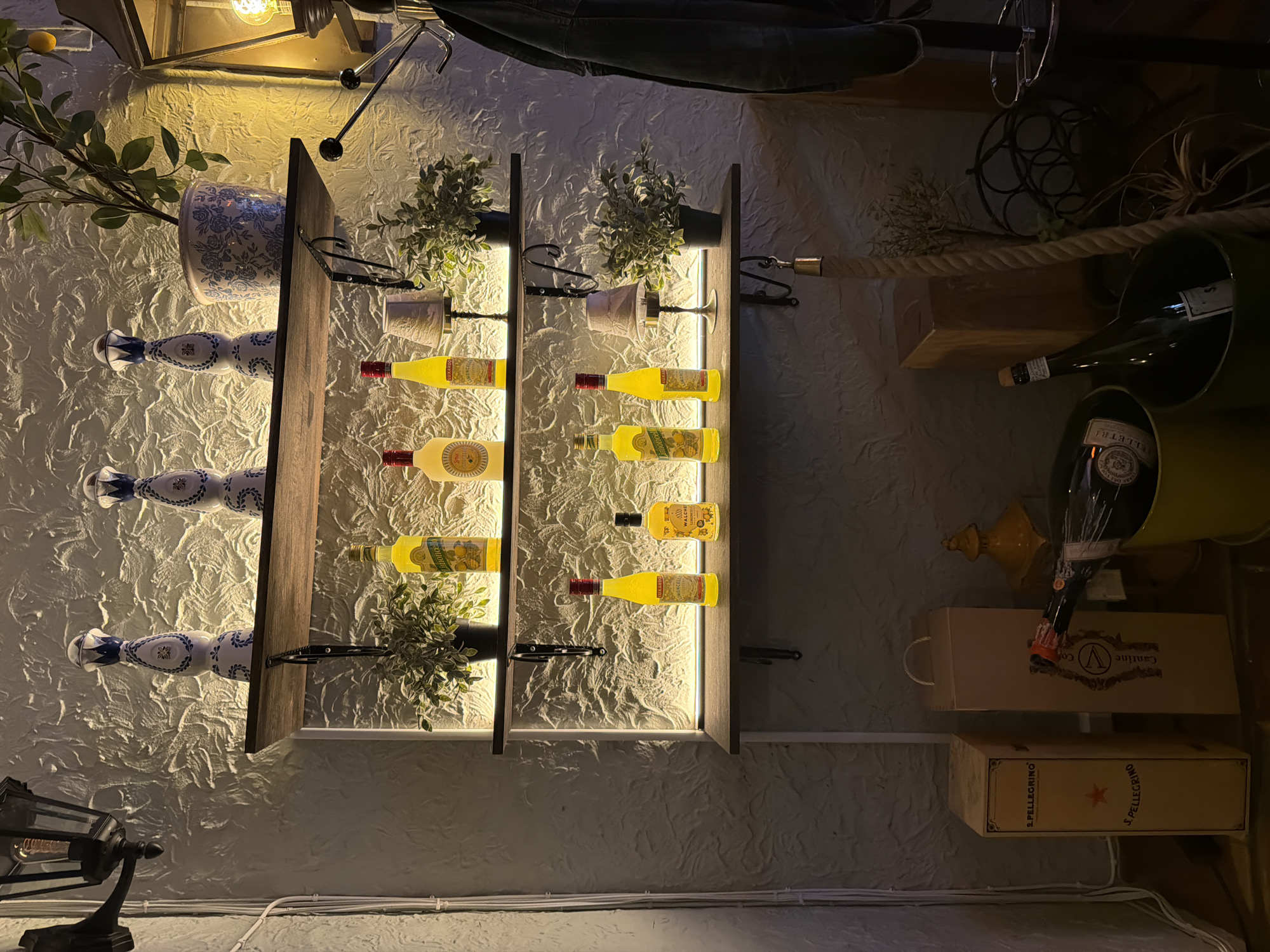 The image shows a dimly lit bar or restaurant interior, possibly in Stockholm, Sweden. The focus is on several shelves displaying various bottles and decorative items. On the top shelf, two decorative bottles with blue and white patterns are visible, alongside a potted plant. The middle shelf holds several yellow bottles, small lamps, and more greenery. The bottom shelf also features yellow bottles, a small lamp, and a potted plant. Below the shelves, several cardboard boxes are stacked, one of which is labeled "S. Pellegrino." A few bottles of wine are also present, with one in a green bucket. A dark jacket is hanging on a rack on the right side of the image. The walls have a textured finish and are illuminated by soft lighting, creating a warm and inviting atmosphere.