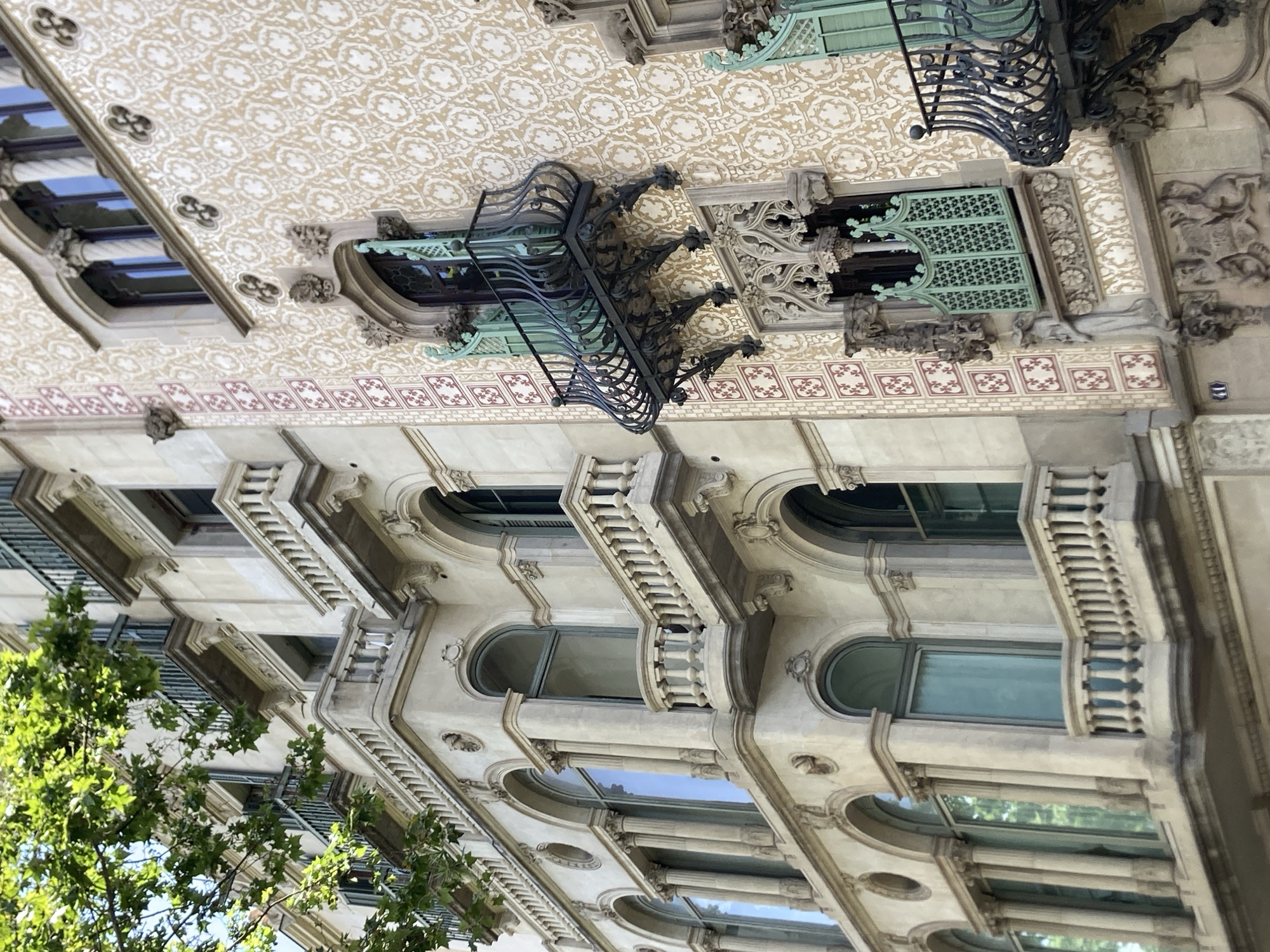 The image shows a detail of an ornate building facade, viewed from below and tilted. The architecture is elaborate, with decorative elements that suggest a historical or Art Nouveau style. The facade features multiple stories with arched windows, intricate wrought-iron balconies, and detailed stone carvings. The upper portion of the building displays a patterned ceiling or frieze with a repeating motif in beige and white. Below this, a band of small, square decorative tiles in red and white is visible. The lower sections of the building are characterized by rows of windows, many of which are arched and framed by decorative stonework. Several balconies with ornate railings are present, adding to the richness of the facade. Lush green foliage from a tree partially obscures the lower left of the building, suggesting it is an outdoor scene. The lighting indicates it is daytime, likely a bright day given the reflections on the windows. No people are visible in the image. The context provided places the location in Barcelona, Spain, a city known for its distinctive modernist architecture.