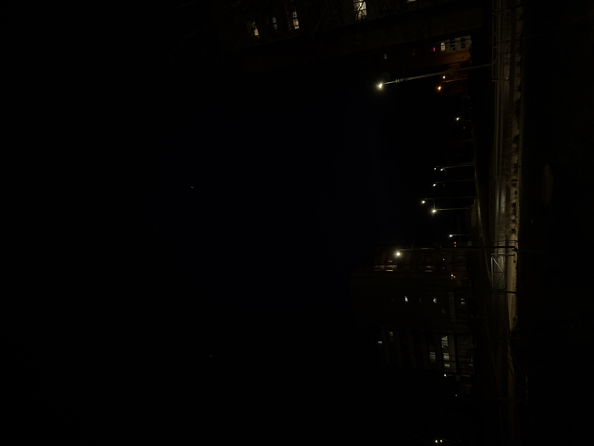The image depicts a night scene in what is likely a city, though specific features are obscured by darkness. Streetlights illuminate a road or path, casting light on what appears to be a wall or structure alongside it. Buildings are visible in the background, with faint lights suggesting windows. The location is unknown, and it is difficult to determine specific characteristics due to the low light conditions. No people or specific actions can be identified in the image. The focus is on the interplay of light and shadow in an urban or suburban setting.