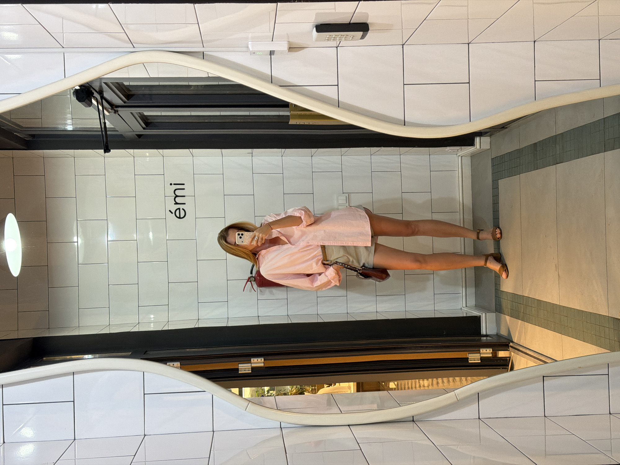 This media file shows a woman taking a mirror selfie in a retail store. She is wearing a light pink oversized shirt and light-colored shorts, paired with brown heeled sandals. She is holding a smartphone in her right hand to capture the photo. A brown purse with a studded strap hangs from her left shoulder. The background consists of white tiled walls and a tiled floor. A fire extinguisher is visible on the wall to her left. The word "émi" is printed in large black letters on the tiled wall behind her. The store appears to be well-lit, suggesting it is daytime or the store is brightly illuminated. The overall ambiance suggests a casual shopping environment.