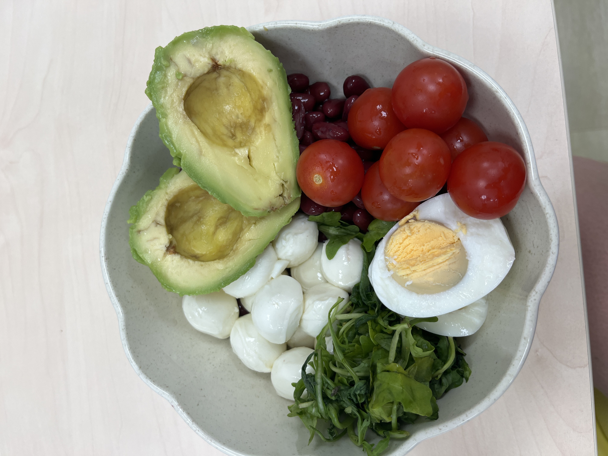 The image shows a close-up, overhead view of a light beige, scalloped-edge bowl filled with a salad. The salad contains several ingredients: two halves of a ripe avocado with visible pits removed, a pile of small, white, round mozzarella balls, a cluster of dark red kidney beans, a portion of green leafy vegetables, several bright red cherry tomatoes, and one hard-boiled egg, halved to reveal its yolk and white. The bowl sits on a light-colored, wood-grain surface. There are no people or visible actions taking place. The image appears to be an indoor shot, likely on a table. There is no information in the image to suggest a location in Plovdiv, Bulgaria.