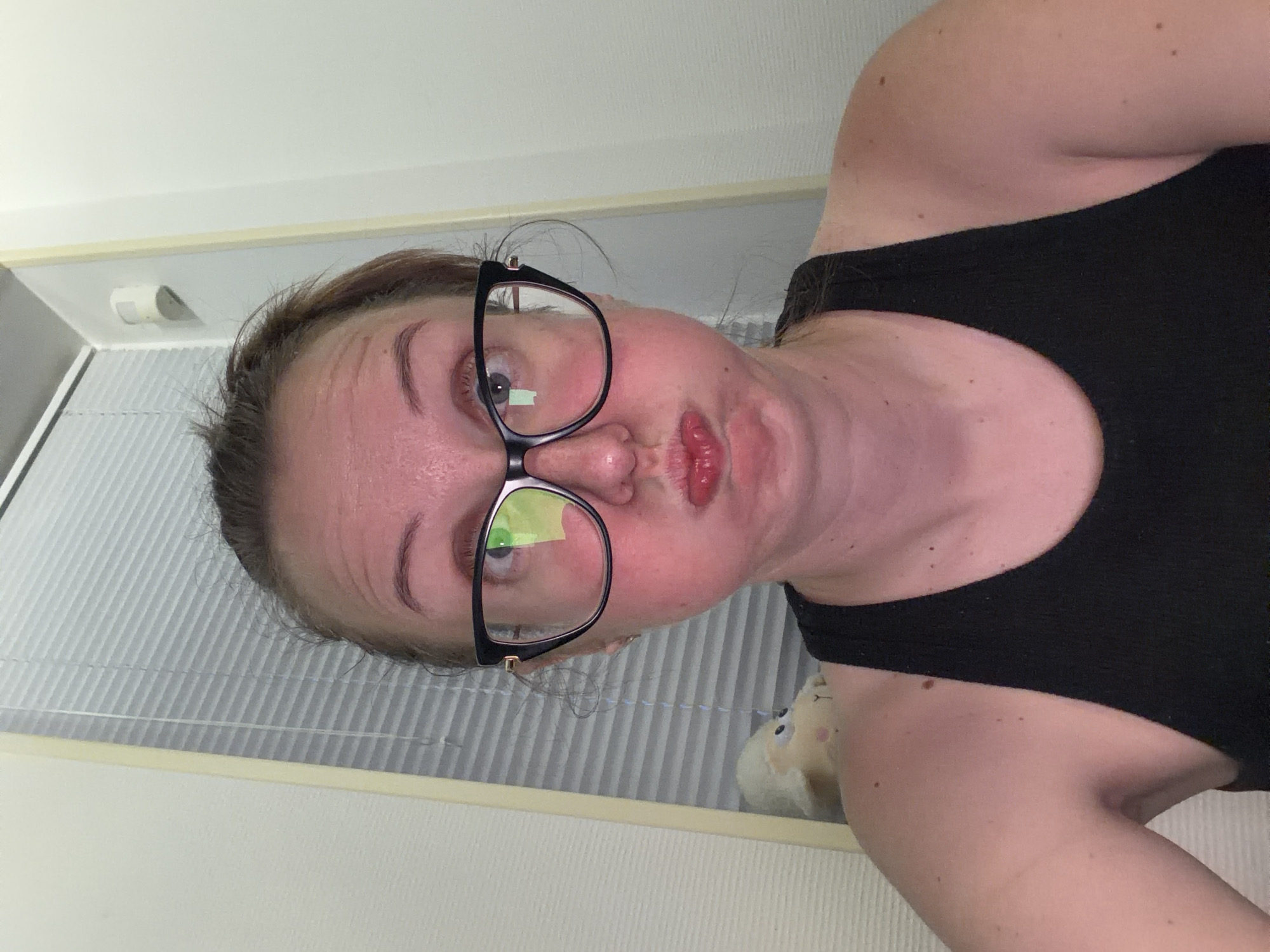 A selfie taken indoors shows a woman with glasses and her cheeks puffed out. She is wearing a black tank top. The background includes a window with blinds and a white wall. A stuffed sheep toy is visible on her left shoulder.