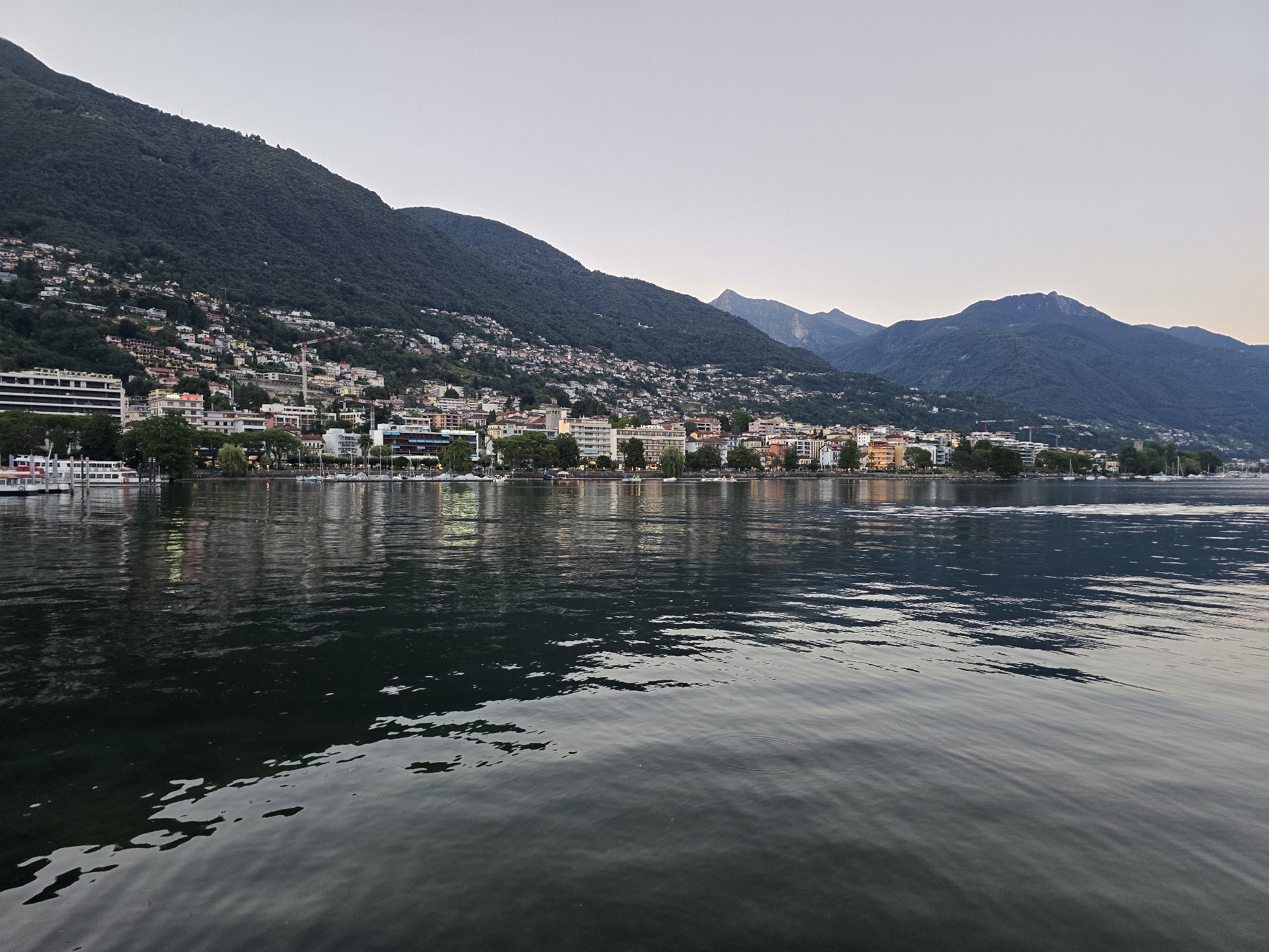 The image depicts a wide, still body of water in the foreground, with the shoreline of a city and mountains in the background. The water reflects the lights from the buildings and boats along the shore, creating a shimmering effect.
The city, which appears to be Locarno, Switzerland, is situated on the slopes of densely forested mountains. Buildings of various sizes and architectural styles line the waterfront, with some appearing to be hotels or apartment complexes. Several boats are docked along the shore, including what looks like a larger ferry or cruise ship on the left side of the image.
The mountains in the background are dark and covered in lush green vegetation. The sky above is a pale, overcast gray, suggesting it might be early morning or late evening. The overall atmosphere is serene and tranquil. There are no visible people in the image, and the scene is devoid of any activity other than the gentle movement of the water. The image captures a picturesque view of a lakeside city nestled against a mountainous backdrop.