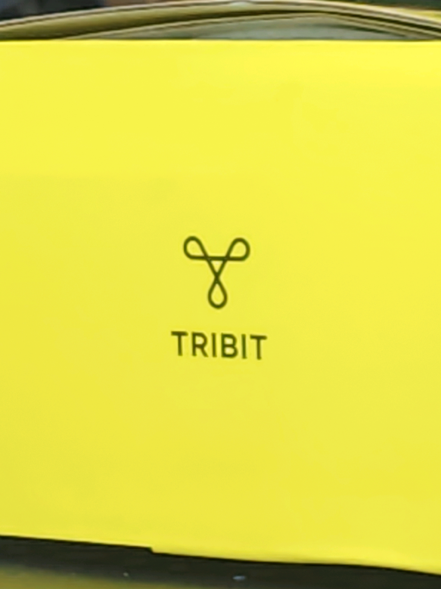 The image shows a yellow box with a black logo and text. The logo appears to be a stylized infinity symbol or a treble clef. Below the logo, the text "TRIBUT" is visible. The box is rectangular and appears to be made of cardboard. The background is blurred but suggests an outdoor setting. The provided crops do not contain any information about the city of Sumerpur, India.