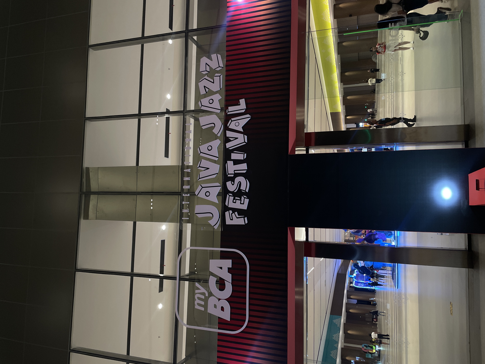 The image shows an interior advertisement for the "Java Jazz Festival" sponsored by "my BCA". The advertisement is displayed on a glass facade of a building, likely a shopping mall or convention center. Through the glass, a view of an interior space is visible, with people walking on a walkway. The location is Salembaran, Indonesia, based on the festival and sponsor. The scene appears to be an indoor public space with ongoing pedestrian traffic.