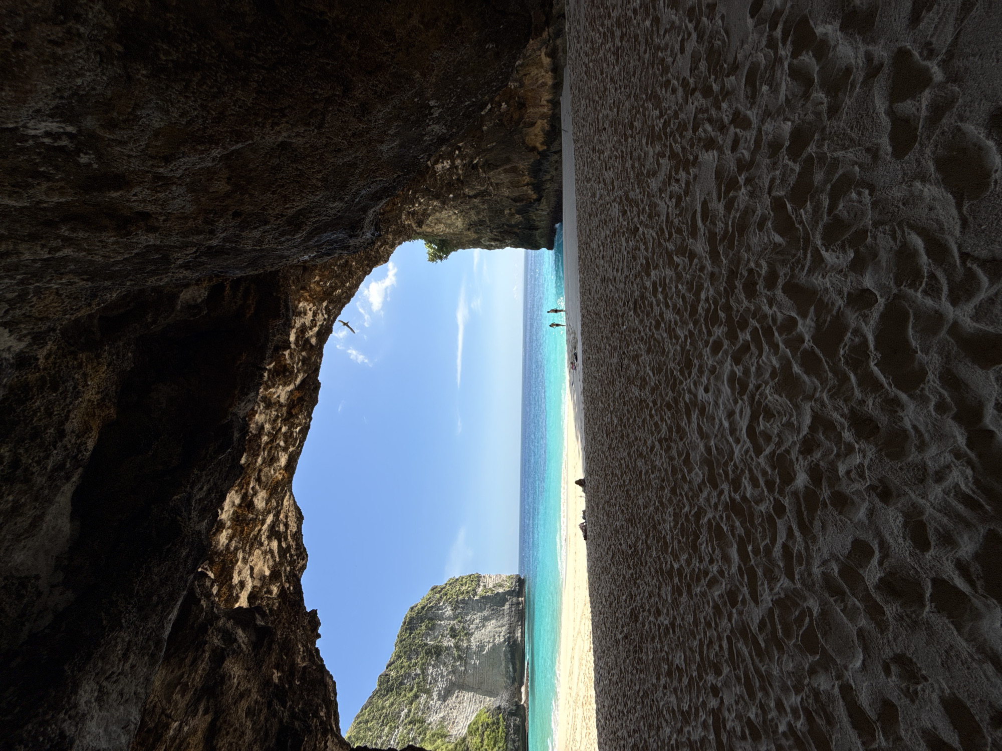 A vertical, medium shot captures a beach scene from within a cavernous opening, likely on the island of Nusa Penida, Indonesia. The perspective is set against a blue sky with a few wispy clouds. The right side of the frame features a sandy beach with patterned, ridged sand extending upwards. The turquoise and azure waters of the ocean form a vertical line between the beach on the right and a jagged rock formation with green foliage on the left. Two indistinct figures stand near the shoreline. The left side of the image is dominated by the dark, shadowed interior of the cave entrance. A single bird flies through the opening.