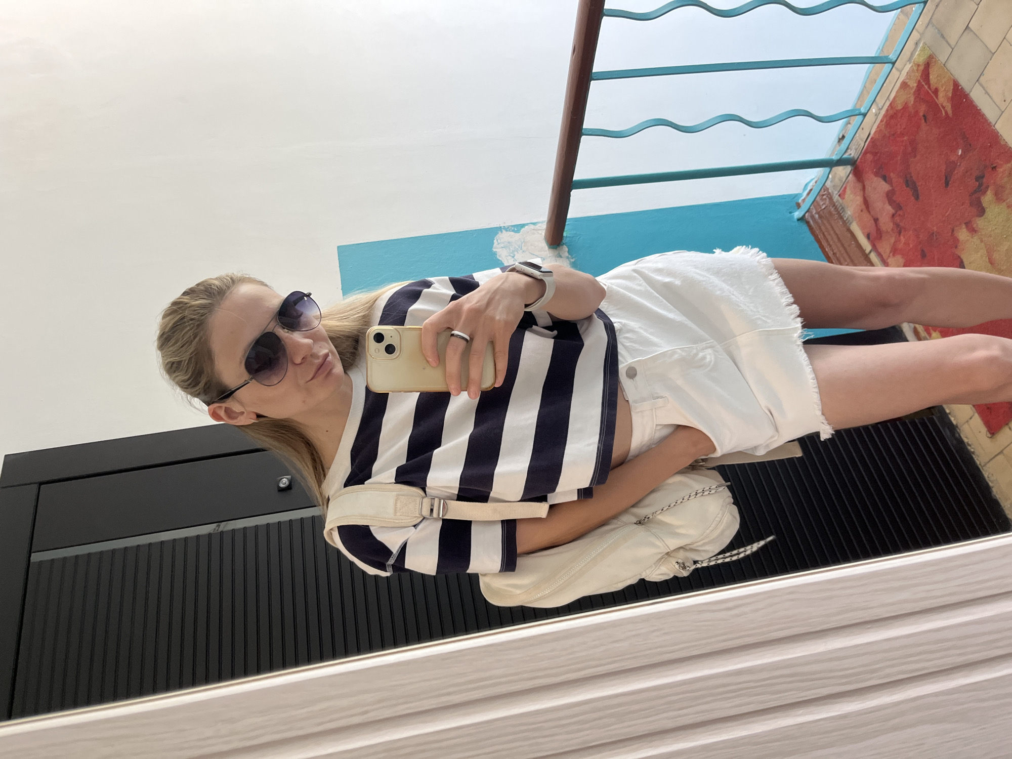 A woman is taking a selfie in a mirror. She is wearing sunglasses, a striped navy and white shirt, white shorts, and a white watch. She has a white bag slung over her shoulder and is holding a phone in her hand. The reflection shows part of a blue railing, a section of tiled floor with red and brown patterns, a white wall, and a black cabinet with vertical lines. The background details hint at an indoor location, possibly a building's interior with a view outside. This is in the city of Chaikovsky, Russia.