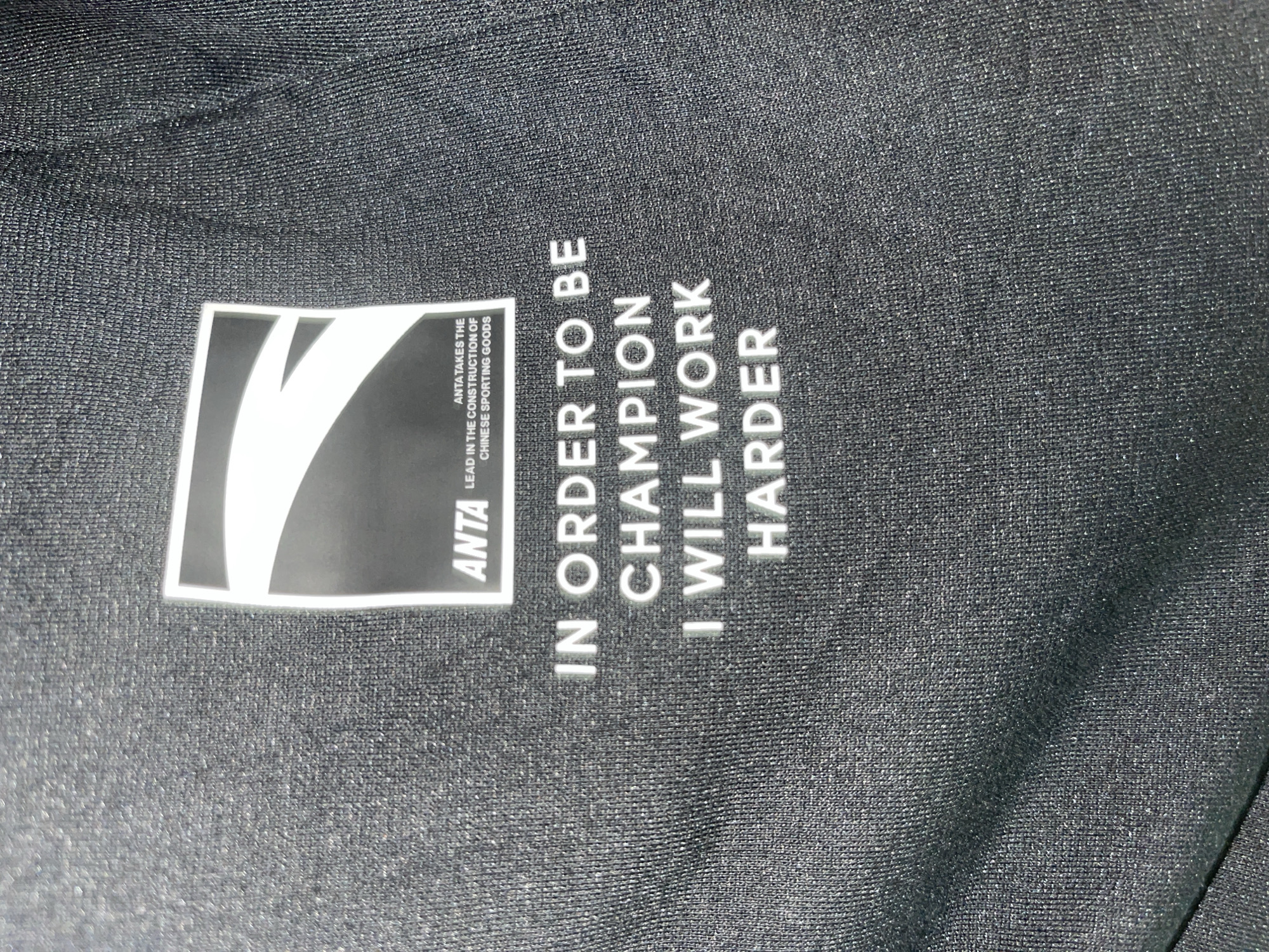 The image displays a close-up, slightly angled shot of a black fabric, likely part of a garment such as sportswear. The fabric exhibits a fine, tightly woven texture with subtle, sparkling flecks that catch the light, suggesting a synthetic material.
Prominently featured on the left side is a rectangular logo in black and white. The logo contains a large, stylized white "V" or wing-like graphic. To its right, the brand name "ANTA" is written vertically in white capital letters. Below "ANTA," smaller text reads: "ANTA TAKES THE LEAD IN THE CONSTRUCTION OF CHINESE SPORTING GOODS."
To the right of the logo, a motivational slogan is printed in white capital letters, oriented vertically. The slogan reads: "IN ORDER TO BE CHAMPION I WILL WORK HARDER."
There are no people, discernible activities, or specific location cues beyond the fabric itself. The lighting appears even, highlighting the printed elements and the fabric's texture. The image focuses entirely on the branding and message on the textile.
