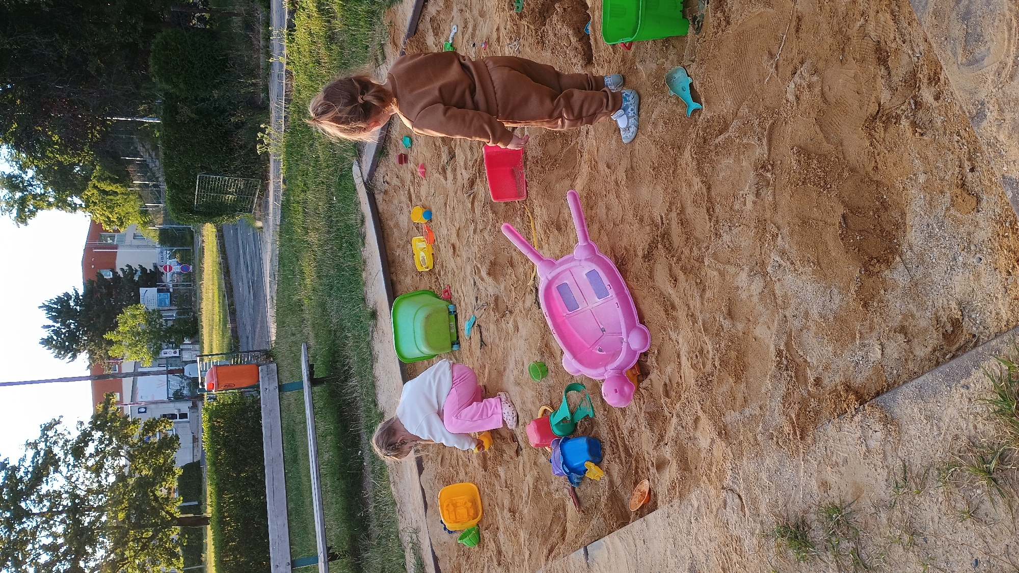 The image depicts a children's sandbox in a park setting, likely in Sokolov, Czechia, given the provided context. Two young girls are present, actively engaged in play. One girl, wearing a white sweater and pink pants, is crouched down and appears to be digging or playing with sand toys. The second girl, dressed in a brown tracksuit, stands facing away from the camera, looking into the sandbox. Various colorful plastic toys are scattered throughout the sand, including buckets, shovels, and a pink toy turtle shell. The sandbox itself is bordered by a wooden frame and surrounded by grass. In the background, a park bench, an orange trash receptacle, and distant buildings are visible, consistent with an urban park environment. The overall scene conveys a sense of outdoor childhood recreation.
