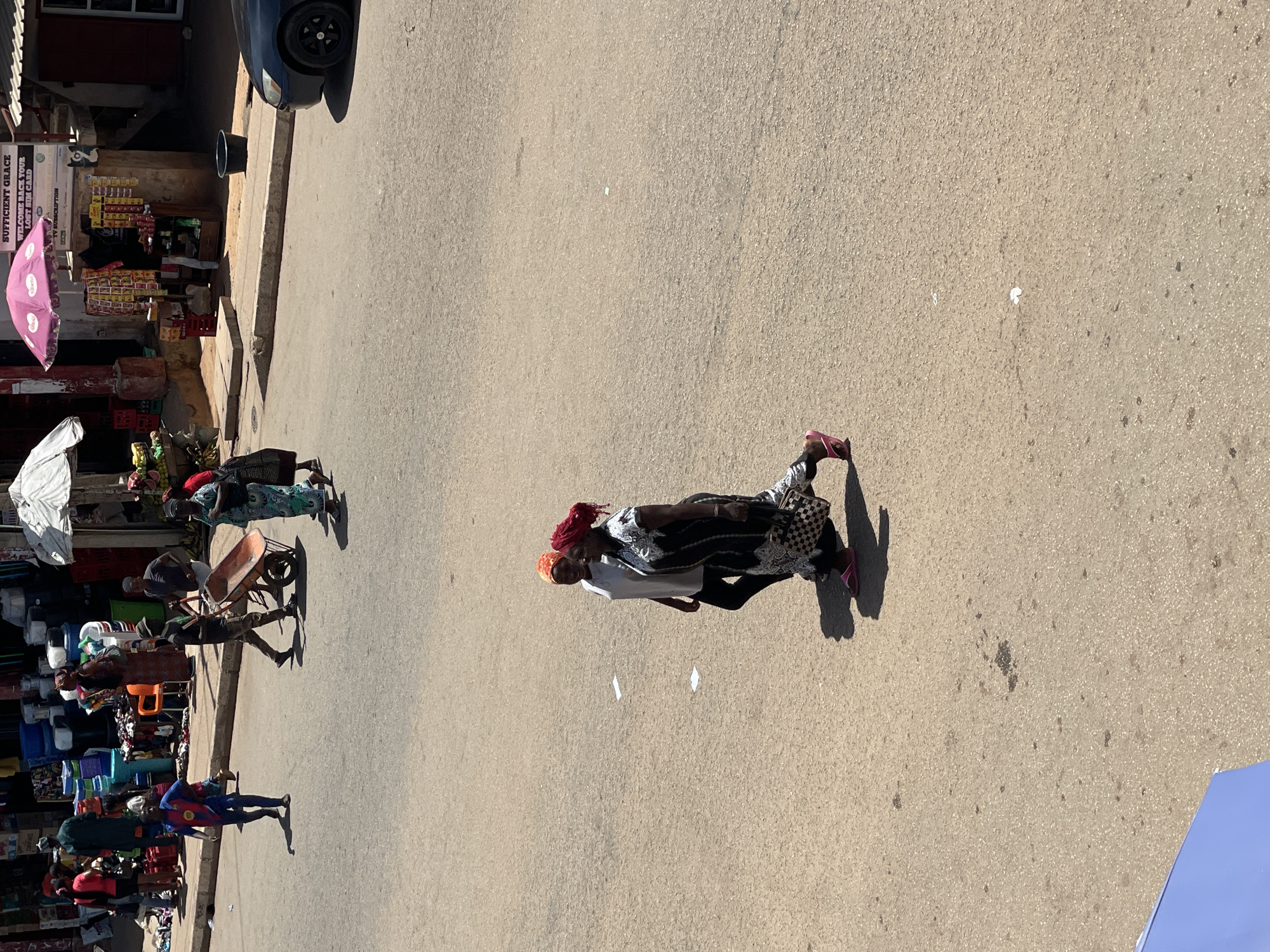 The image depicts a street in Bukuru, Nigeria, with various people and objects visible. On the left side of the frame, there are shops displaying assorted goods, including buckets, clothing, and groceries under umbrellas. People are seen walking and interacting with the shops. In the center, two women are walking along the street. One woman has a red head covering, a white shirt and black leggings. The other woman has a red head covering and is wearing a patterned dress. The street itself is paved. A parked car is visible in the upper left corner.