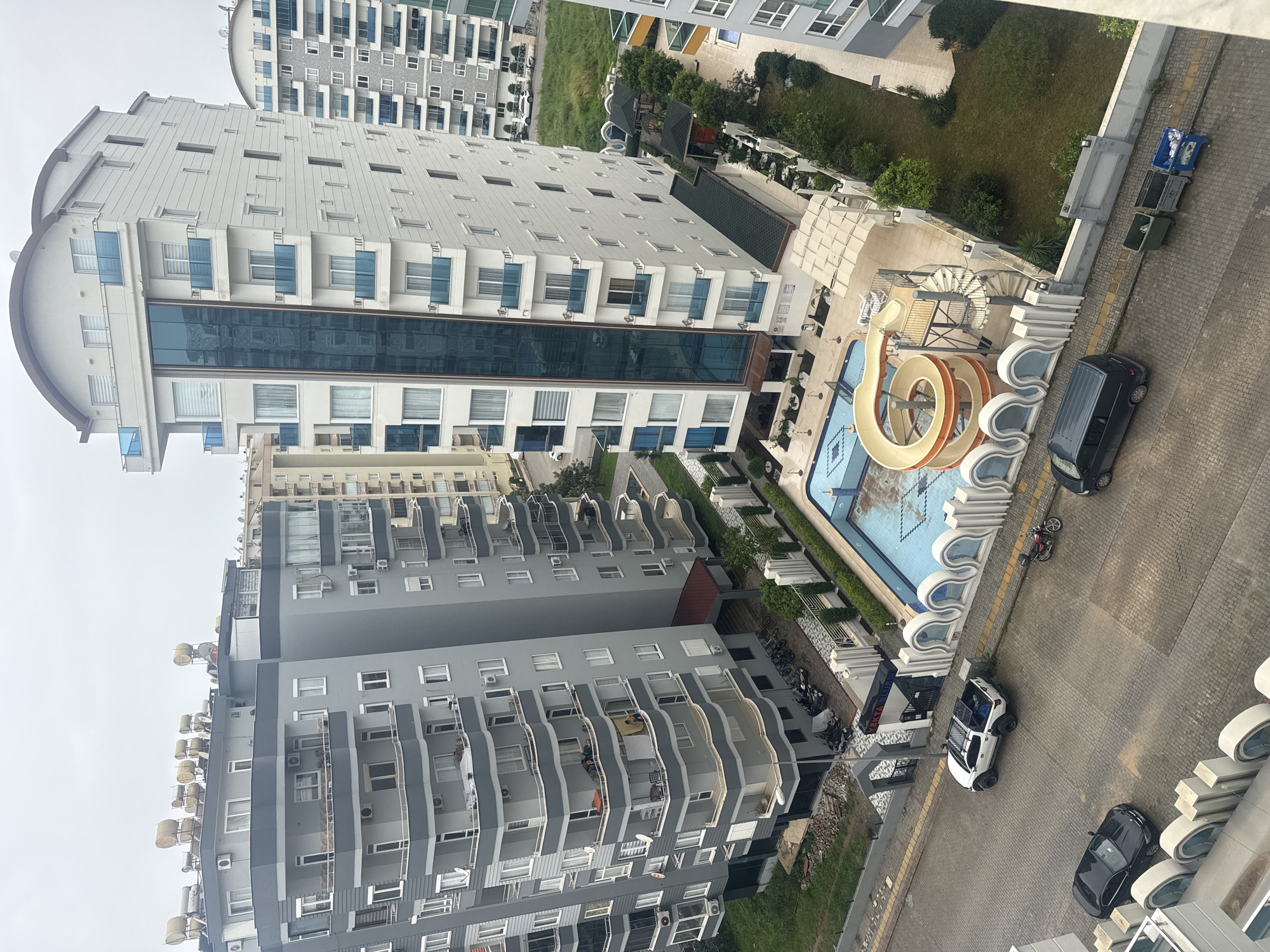 The image shows a view from above of a cityscape in an unknown city. The scene includes several high-rise buildings, with two prominent structures in shades of gray and white with blue accents. The gray building has balconies with various items on them, including what appears to be furniture and laundry. The white building has a curved top and a central section of blue glass.
Between the buildings is a swimming pool with a water slide, although the pool appears empty. Adjacent to the pool, there's a walkway and some landscaping. A few vehicles are parked on a paved surface beside the buildings, including a white vehicle with an open door and a black van.
In the background, more buildings can be seen, partially obscured by greenery. Along the edge of the paved area, there are what seem to be garbage bins or recycling containers. The overall atmosphere suggests an urban or suburban residential area.