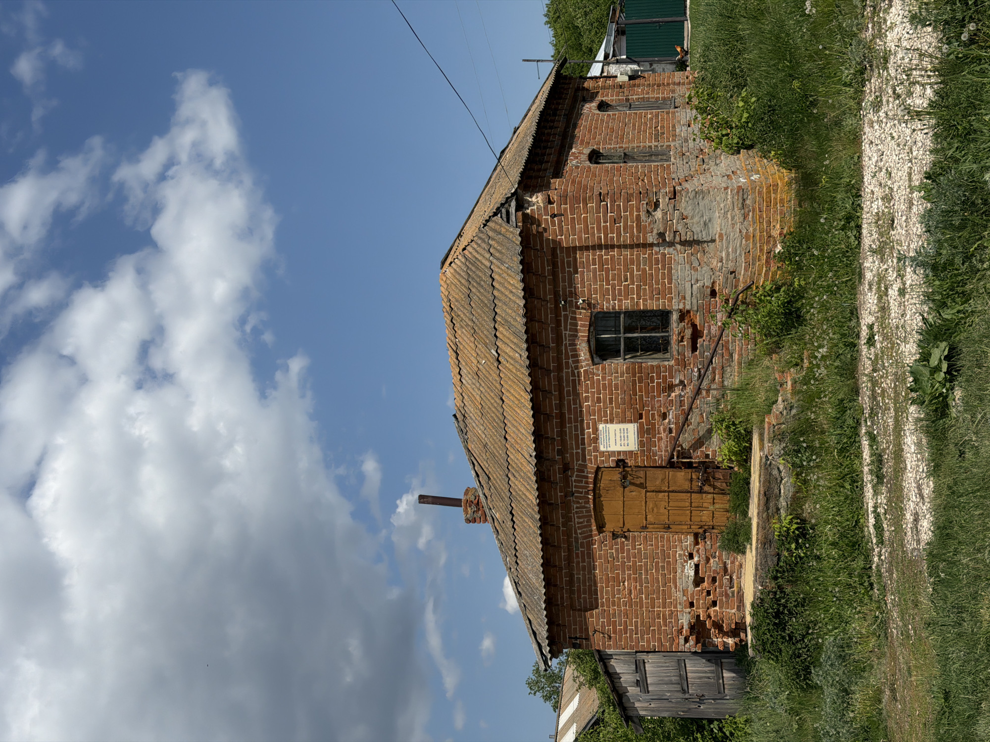 The image depicts a two-story brick building in Полянки, Russia. The building has a weathered, brown roof and a small chimney. The brick facade shows signs of disrepair with missing bricks. A wooden door is visible on the first floor, along with a sign. Windows are seen on both floors, some with ornamental grilles. The surrounding environment includes a gravel path, grass, and foliage, with a cloudy sky overhead.