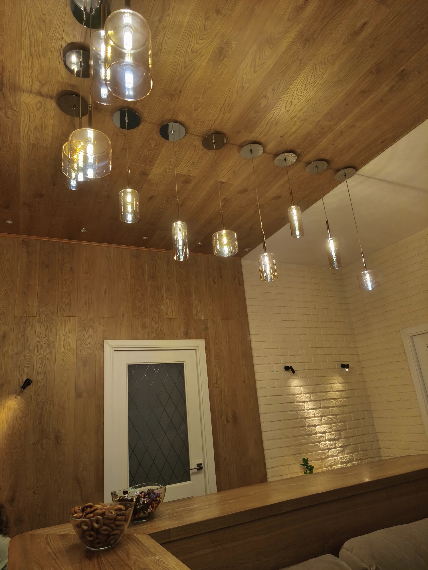 This image depicts an interior scene, likely a residential or commercial space, with a focus on the ceiling and upper wall areas. The ceiling is finished with wood paneling, displaying a warm, natural wood grain pattern. Hanging from the ceiling are multiple pendant lights. Some are clusters of cylindrical glass shades, while others are single, elongated glass fixtures. These lights emit a warm, yellowish glow. The upper portion of the right wall is covered in a textured white brick finish, and two small, black directional spotlights are mounted on it, casting light downwards. The lower portion of the left wall and a door are paneled with the same wood as the ceiling. A white door with a diamond-patterned glass insert is visible in the wood-paneled section of the wall. In the foreground, a wooden counter-like surface holds two glass bowls; one contains numerous small, ring-shaped baked goods, and the other contains assorted candies. The overall impression is of a modern and warmly lit interior. There are no people or animals visible in the image. The location is not specified in the image, and therefore, it cannot be confirmed to be in Vladivostok, Russia.