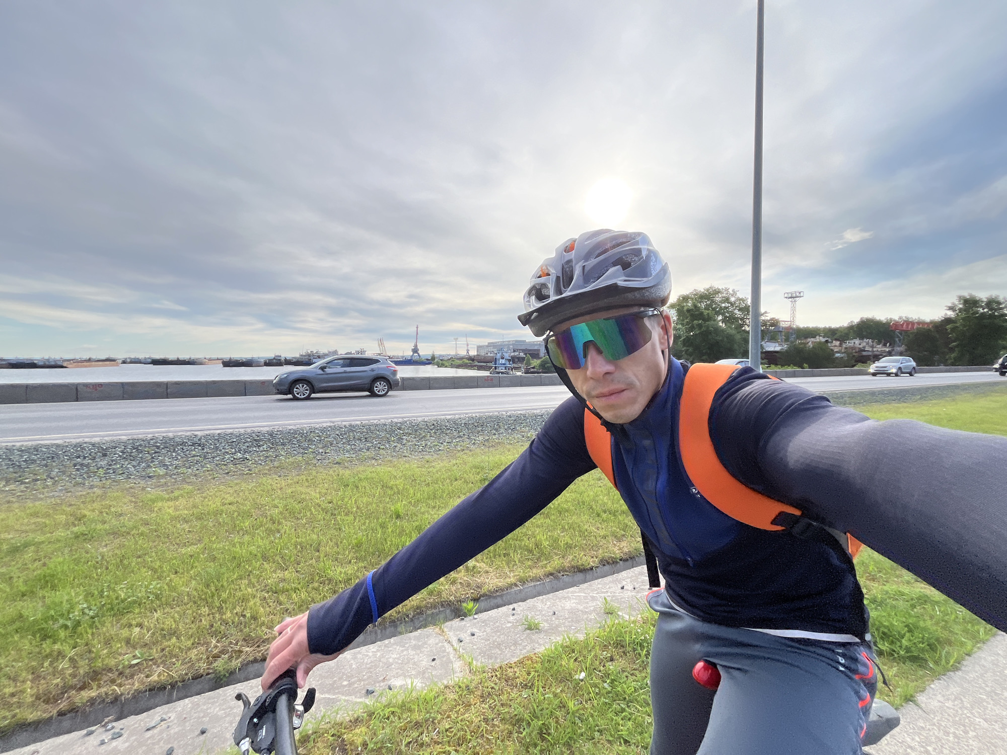 A man on a bicycle takes a selfie on a road near Chaikovsky, Russia. He wears a helmet, sunglasses, a long-sleeved cycling jersey, and dark trousers. He is positioned on the side of the road, next to a patch of grass and a low concrete barrier. Behind him, a road is visible with a gray car driving on it. Beyond the road, there is water, with some industrial structures visible in the distance. In the background, the sky is overcast, and the sun is shining through the clouds. A lamp post stands upright near the road.