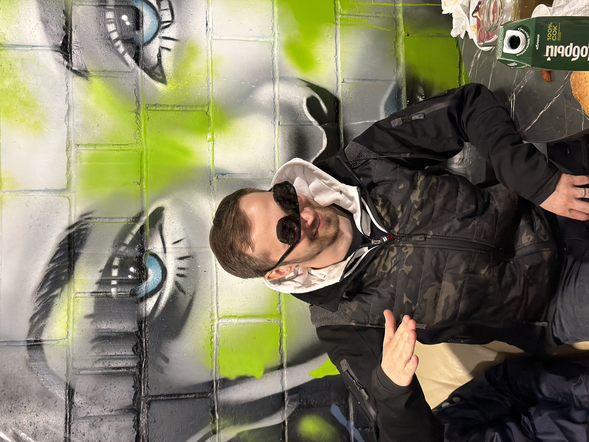 A man wearing sunglasses, a camouflage jacket over a white hooded sweatshirt, and dark pants is seated, gesturing with his left hand. A table is partially visible to the right, holding a box of juice, a glass, and a partially visible pastry. Behind the man is a wall mural depicting a large face with prominent eyes painted on what appears to be a brick surface. The juice box has text in Russian, including "100% COK". The location is possibly Ust-Ilimsk, Russia based on the juice box.