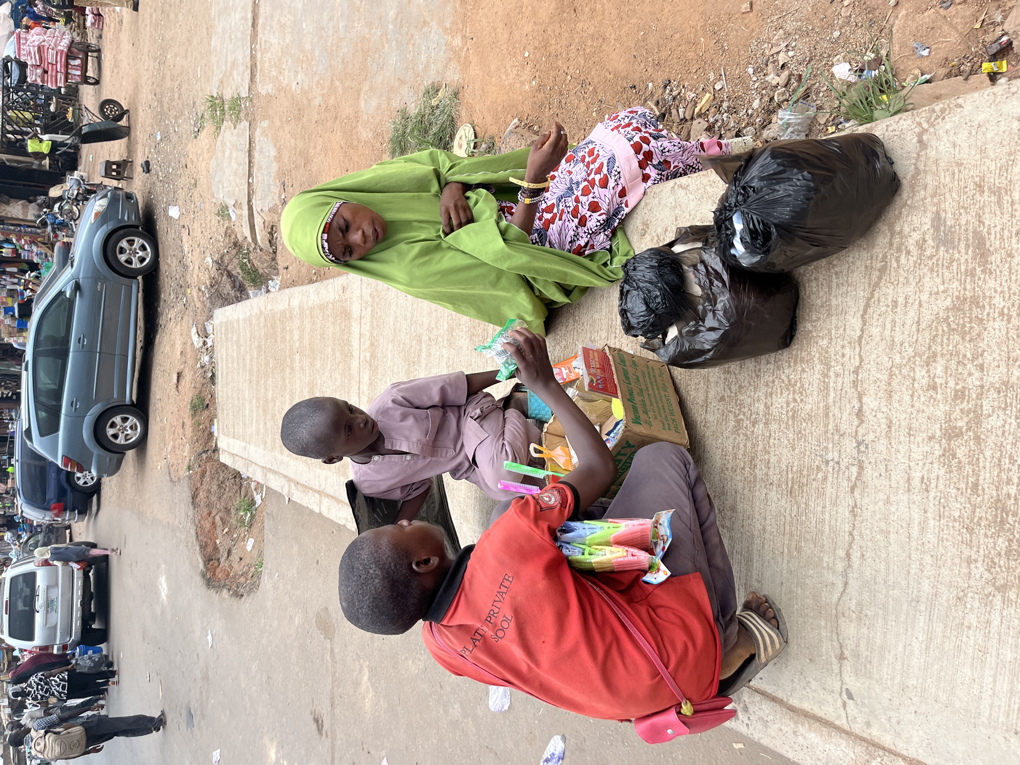 Here is a description of the image:
A medium, eye-level shot of a scene taking place in the city of Jos, Nigeria. Three people are sitting on a concrete ledge next to a road. One person, wearing a green headscarf and a floral patterned dress, is reclined. Two other children, one wearing a red shirt that says “PLATE PRIVATE SOOL” are sitting upright. The child in red has a collection of colorful plastic objects in his lap. There are two black plastic garbage bags next to the reclined person. The ledge is next to a road with cars and people walking. A green cardboard box sits near the child in red. The background shows a busy market street with multiple vehicles and pedestrians.