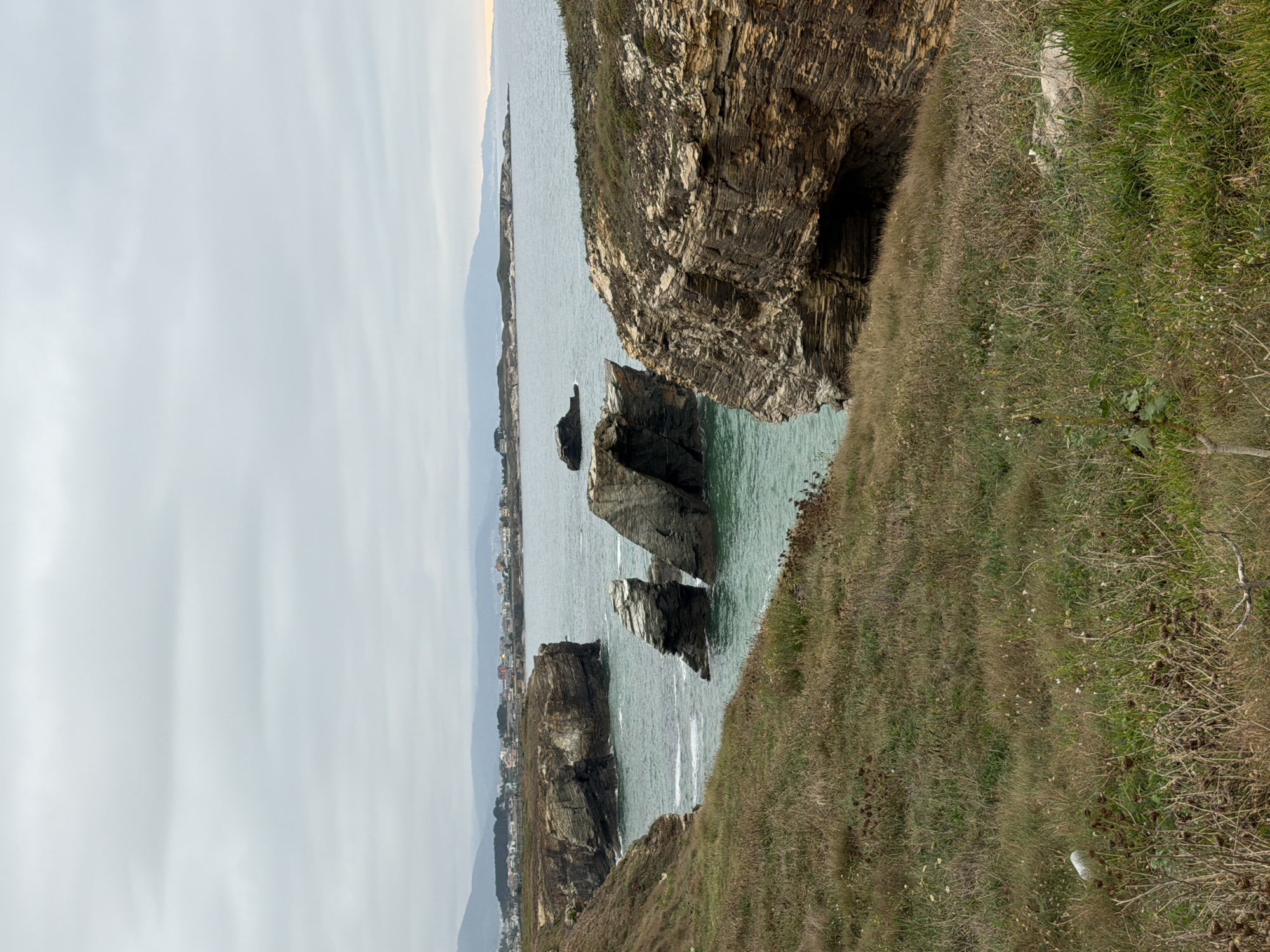 The image depicts a coastal landscape in Ribadeo, Spain. The foreground is dominated by a grassy slope with dry, brown vegetation and some scattered green shoots. A white object, possibly litter, is visible on the grass in the lower left quadrant. The middle ground features a rugged, layered rock formation jutting out from the cliffside, with a dark opening resembling a cave or overhang. Beyond this, the turquoise water of the sea is visible, with waves lapping against the shore. In the distance, several large, weathered rock arches and sea stacks rise from the water, some with vegetation clinging to their tops. The background shows a coastline with buildings of a town nestled along the hills, and further out, a hazy mountain range under an overcast sky. The overall impression is of a wild, natural coastal environment.