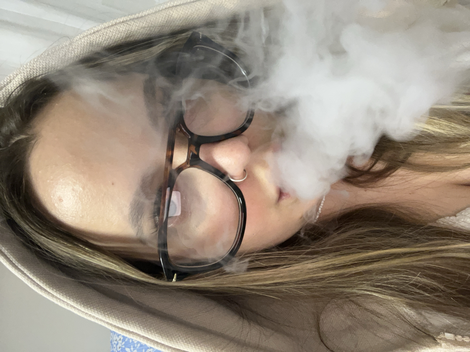 This close-up image, oriented at an angle, features a person's face largely enveloped by a dense cloud of white vapor or smoke. The individual wears glasses with dark, possibly tortoiseshell, frames that partially obscure their eyes. A silver nose ring is visible on their right nostril. Light brown hair with blonde highlights frames their face, and they appear to be wearing a light-colored hooded garment, possibly a sweatshirt or jacket, which is visible around the top and bottom edges of the frame. The background is indistinct and appears to be a plain, light surface, suggesting an indoor setting. The primary activity is the exhalation of a large volume of vapor, indicating vaping or smoking. Due to the smoke and the angle, the person's expression and specific emotions are not clearly discernible. There is no visible text in the image.