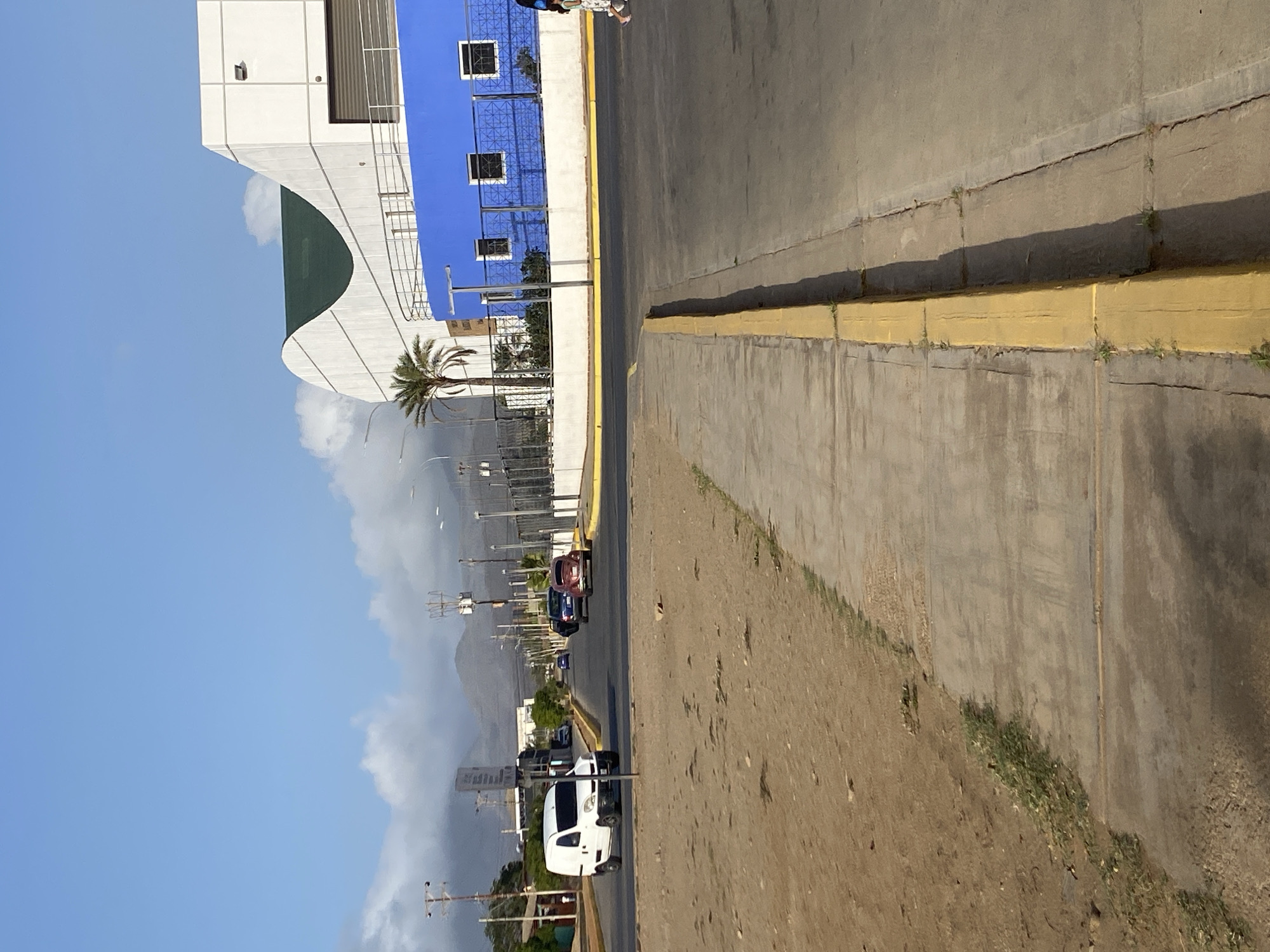 The image shows a street scene, likely in Juan Griego, Venezuela, featuring a building with white and blue sections, a curved green awning, and a palm tree. The street runs parallel to the building and is lined with yellow curbs. Several cars are visible on the street, including a white van in the foreground. In the distance, mountains are visible under a cloudy sky. The perspective is slightly angled.