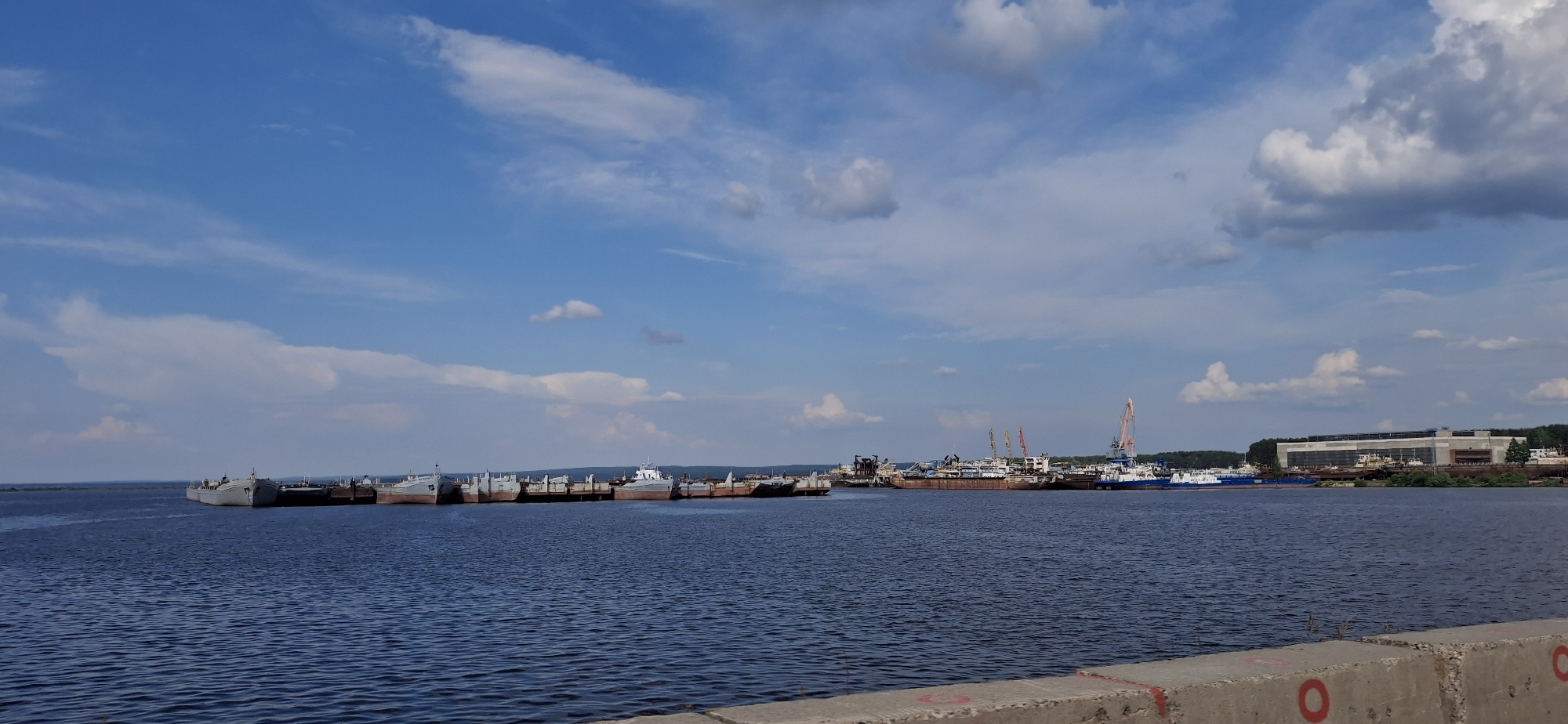 A wide, panoramic, eye-level, outdoor, daylight, horizontal medium shot captures a vast blue sky with scattered white clouds above a wide, calm, dark blue body of water. Along the far bank, a line of various ships and barges are docked. Several large cargo ships, some with red hulls, are visible, as are some smaller vessels and barges. Prominent cranes, one pink and several other smaller ones, rise above the ships, indicating a port or shipyard. In the distance, a large, modern building with a light-colored facade and dark roof is situated along the tree-lined shore. The foreground features the edge of a concrete structure, likely a pier or embankment, with red painted circular markings. The scene appears to be in the city of Chaikovsky, Russia, likely at a port or industrial area on the Kama River.